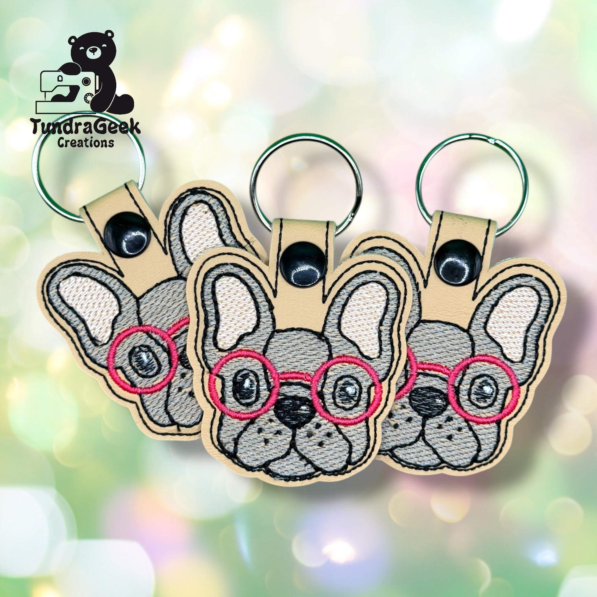 Three embroidered keychains featuring a French bulldog face with red glasses are arranged side by side on a light bokeh background. Logo TundraGeek Creations is in the top left corner.