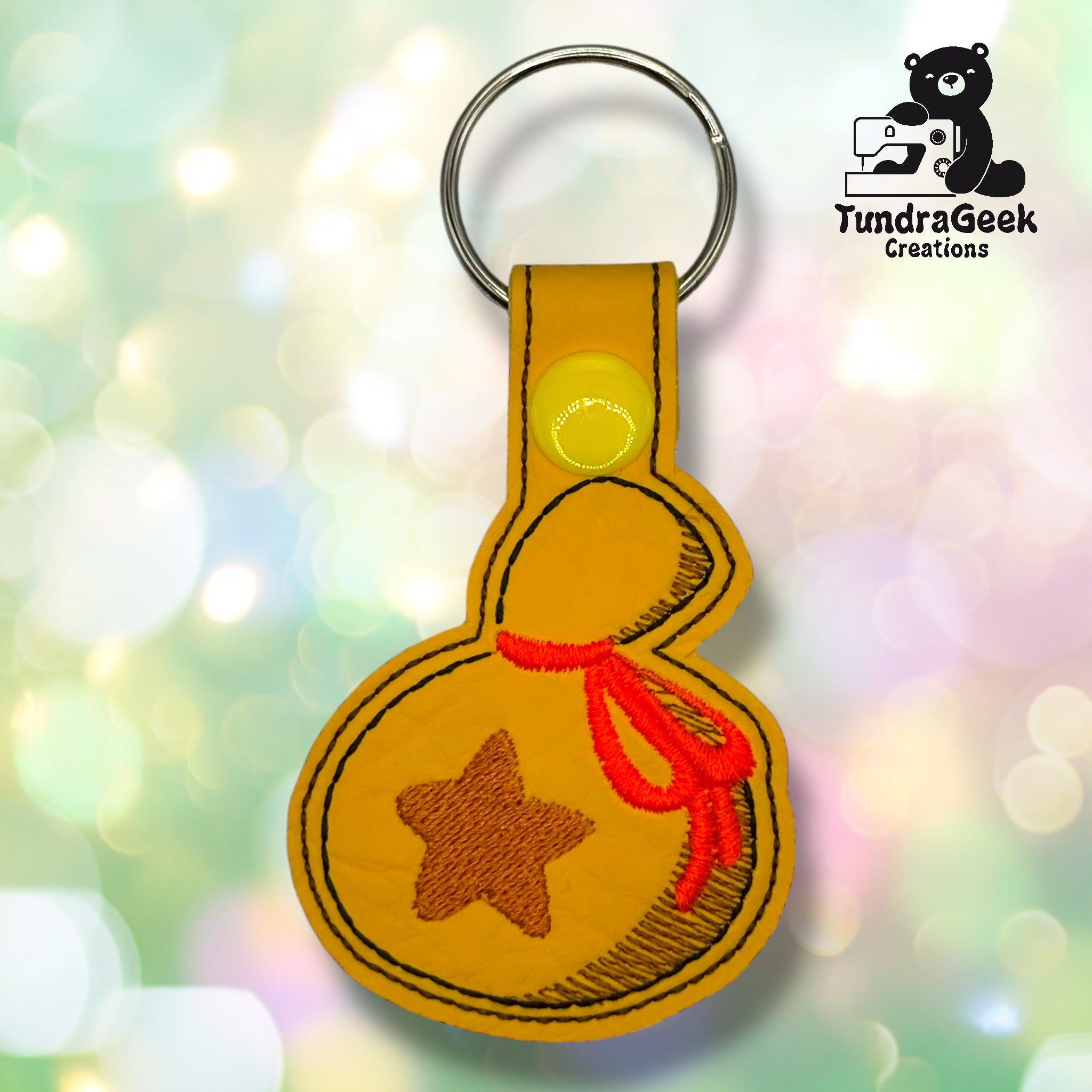A yellow keychain in the shape of a money bag with a brown star, red string detail, and black stitching, displayed with the TundraGeek Creations logo in the corner.