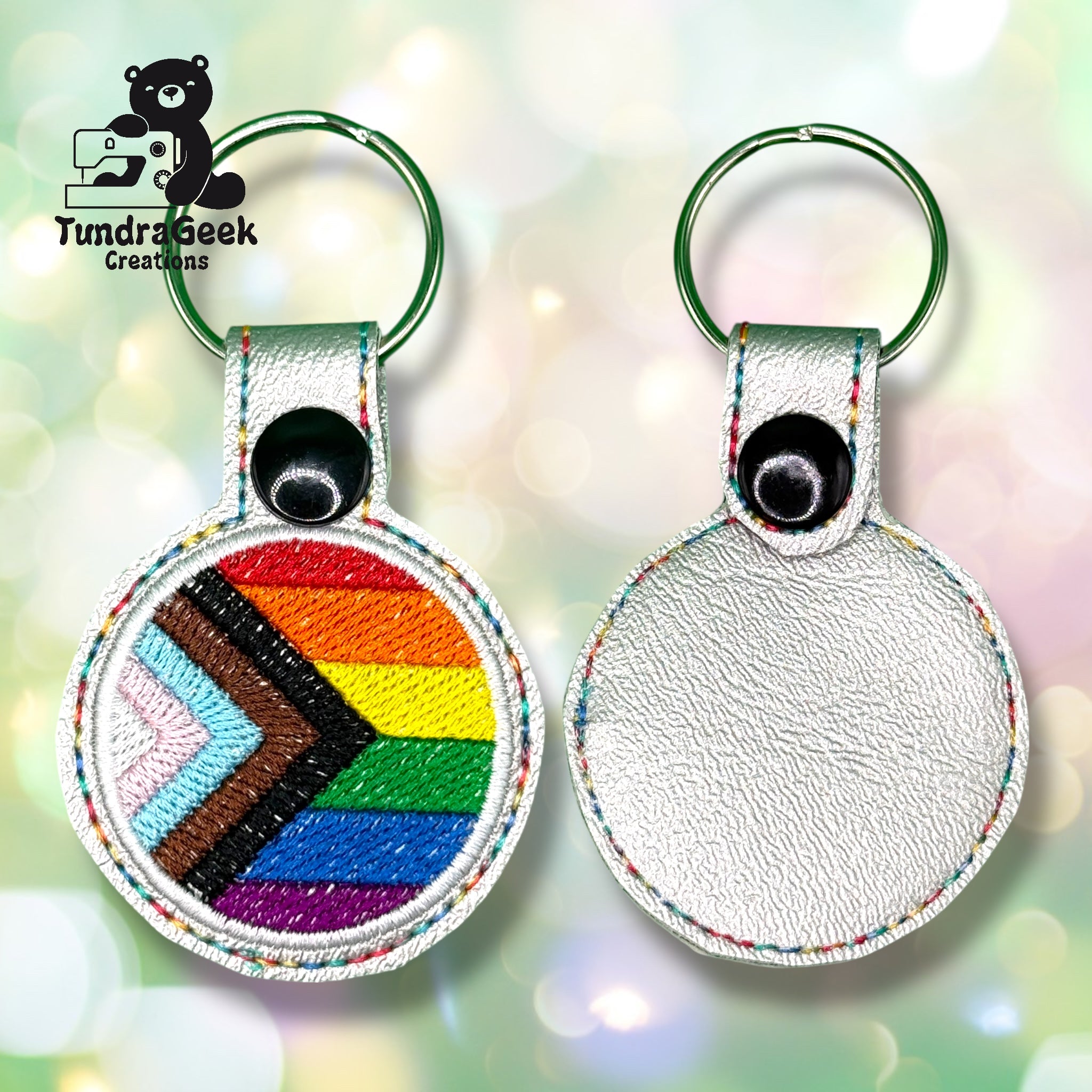Two circular keychains made of silver-colored material, one showing the Progress Pride Flag design, the other blank; both have metal key rings and a snap button at the top.
