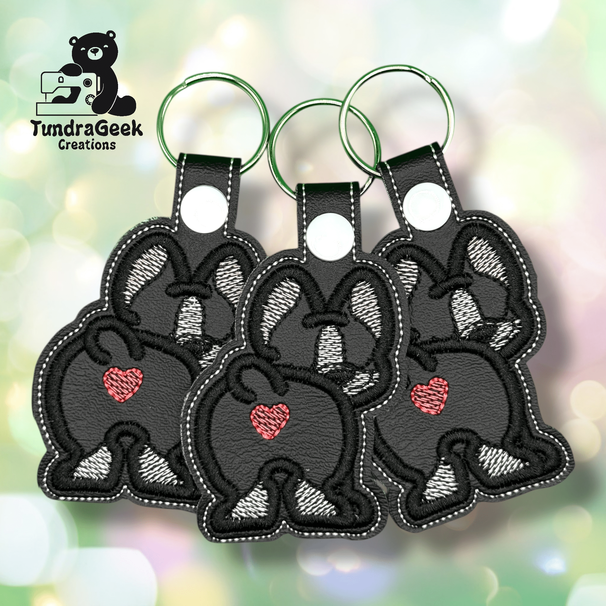Three black embroidered keychains shaped like bunny backsides, each with a pink heart and a green keyring, are displayed on a soft, pastel bokeh background.