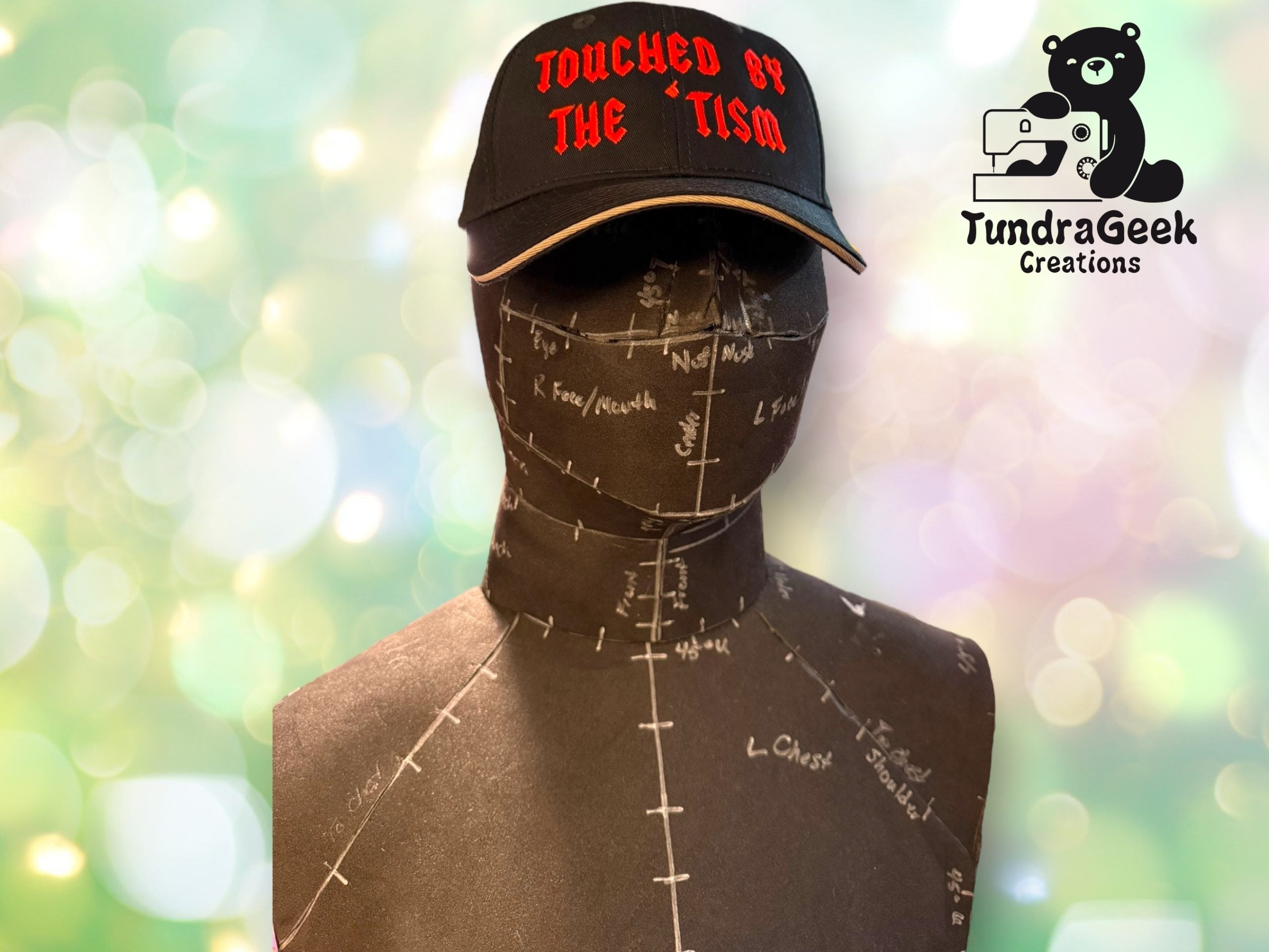 A mannequin wearing a black cap with red text reading TOUCHED BY THE TISM. The mannequin is marked with measurement lines; the TundraGeek Creations logo is in the upper right corner.