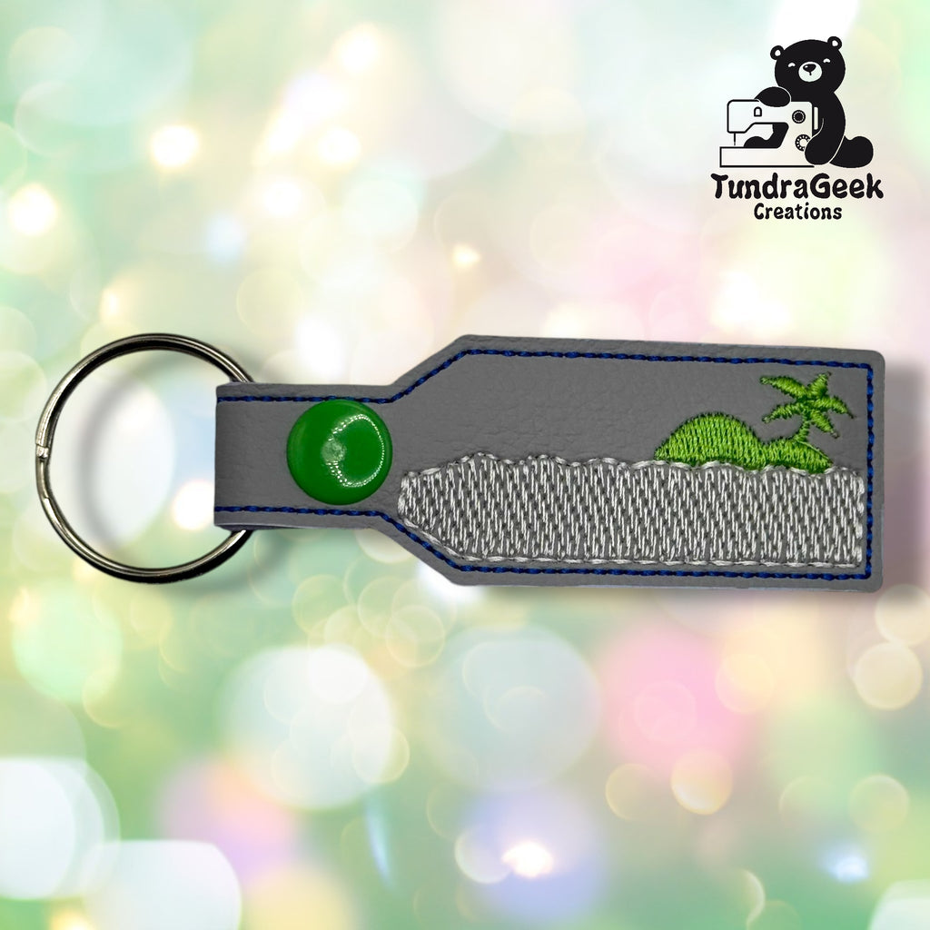A rectangular grey keychain with a green snap button features a stitched design of a green dinosaur and palm tree. The TundraGeek Creations logo is in the top right corner.