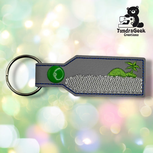 A rectangular grey keychain with a green snap button features a stitched design of a green dinosaur and palm tree. The TundraGeek Creations logo is in the top right corner.