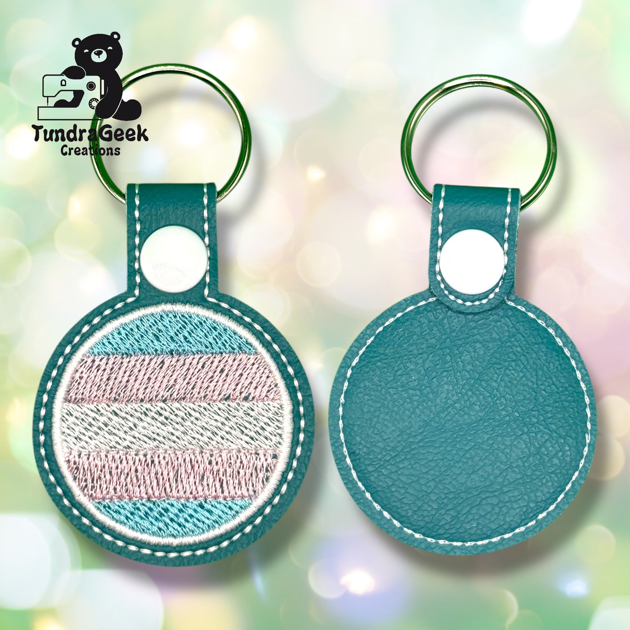 Front and back views of a teal-round keychain with light stitching and a pastel-striped embroidered section on one side. Tundra Geek Creations logo in the upper left.