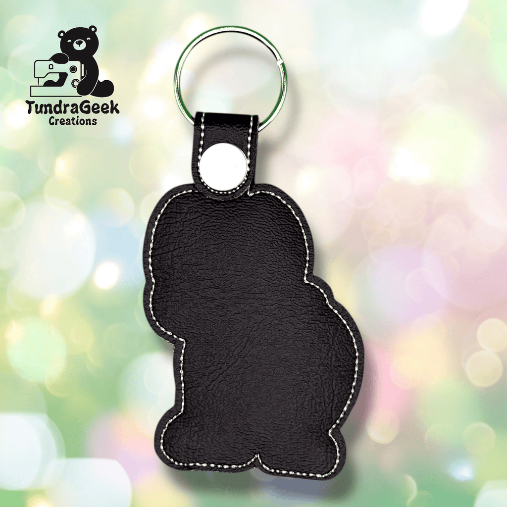A black faux leather keychain in the shape of a seated bear, with visible stitching and a metal keyring, displayed against a colorful blurred background.