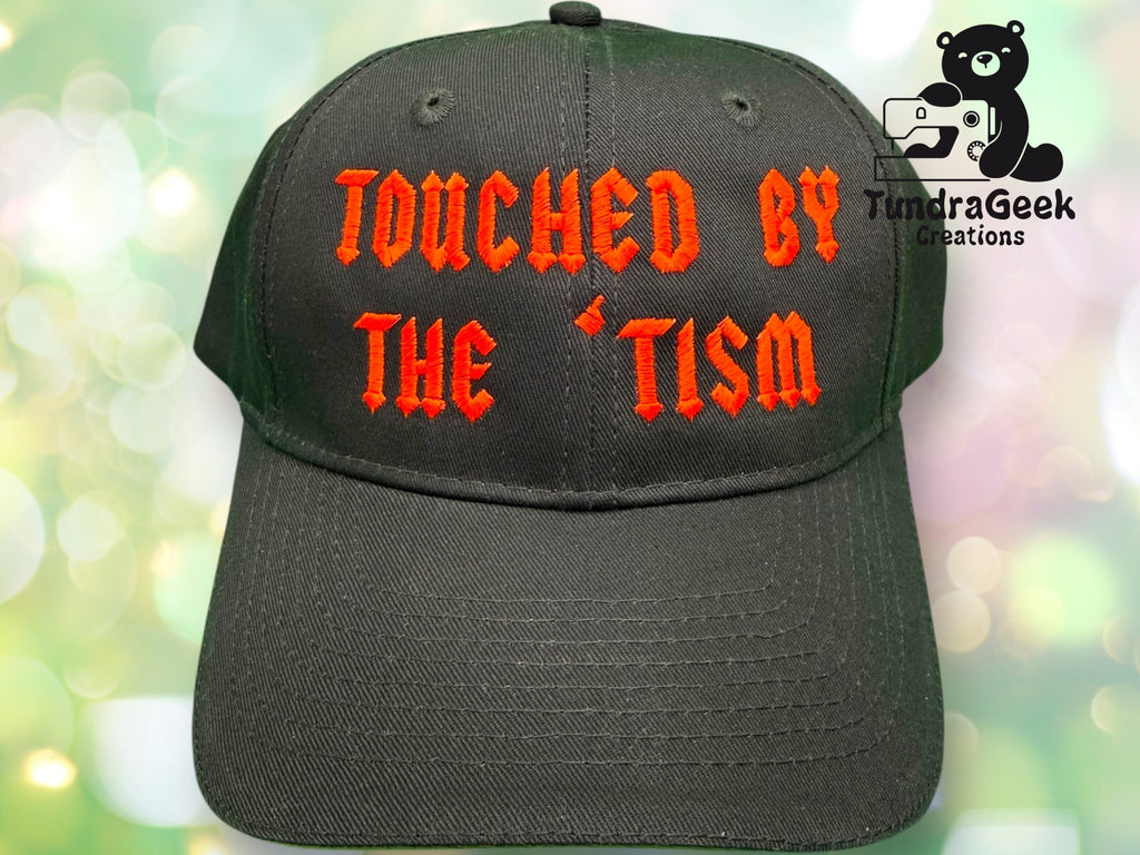 A black baseball cap with red gothic text reading TOUCHED BY THE TISM and a logo for TundraGeek Creations in the background.
