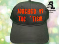 A black baseball cap with red gothic text reading TOUCHED BY THE TISM and a logo for TundraGeek Creations in the background.