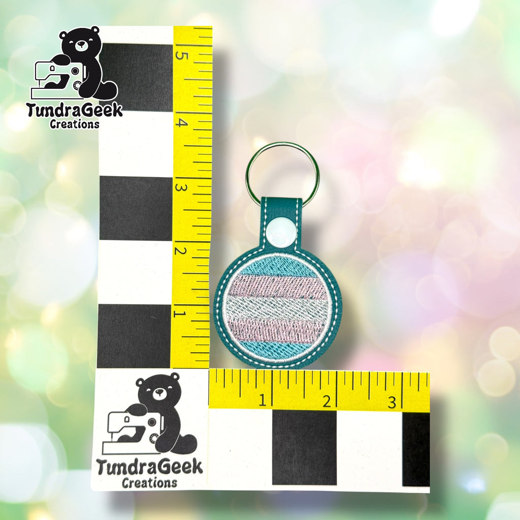 A round keychain with pastel striped embroidery is displayed upright against a measuring ruler with TundraGeek Creations branding.
