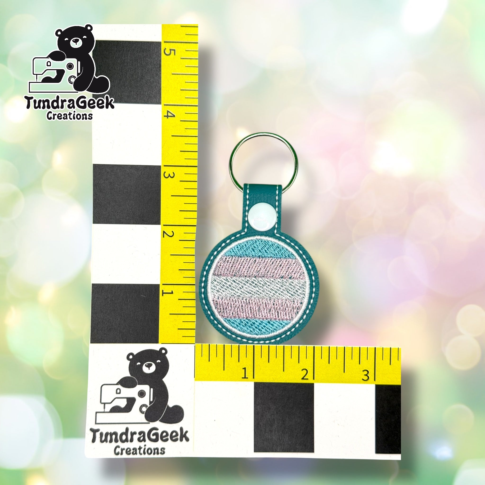 A round keychain with pastel striped embroidery is displayed upright against a measuring ruler with TundraGeek Creations branding.