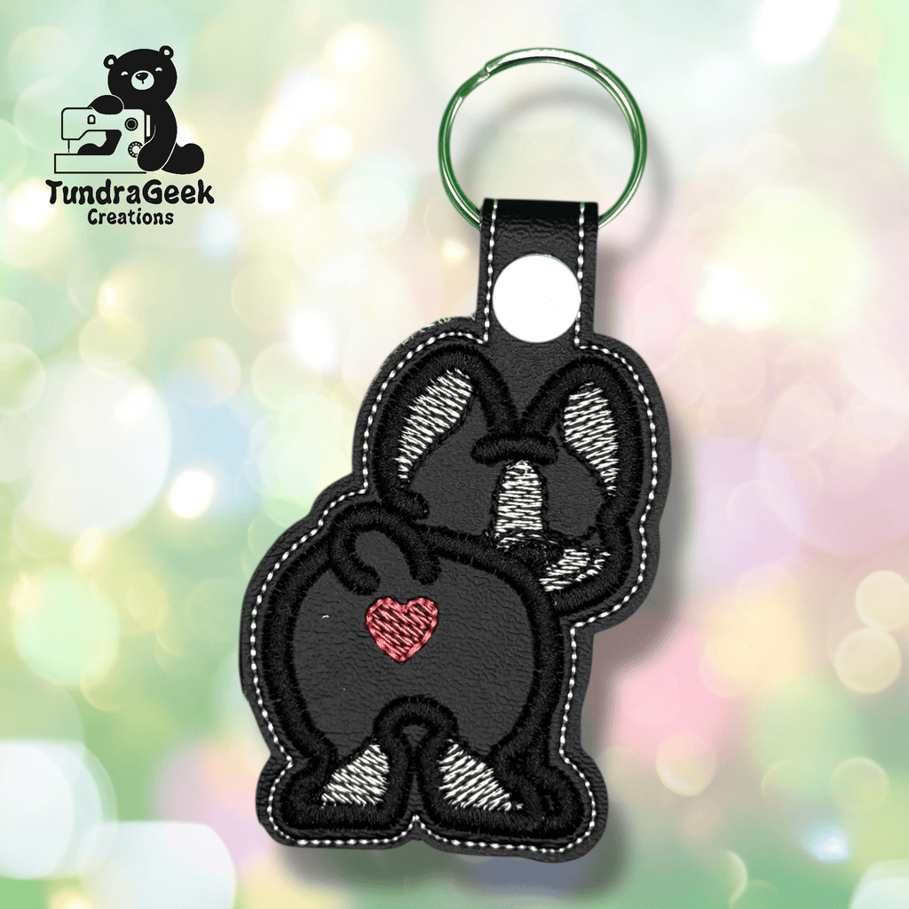 A keychain featuring a black and white embroidered bunny with a red heart on its rear, set against a blurred pastel background. TundraGeek Creations logo appears in the top left corner.