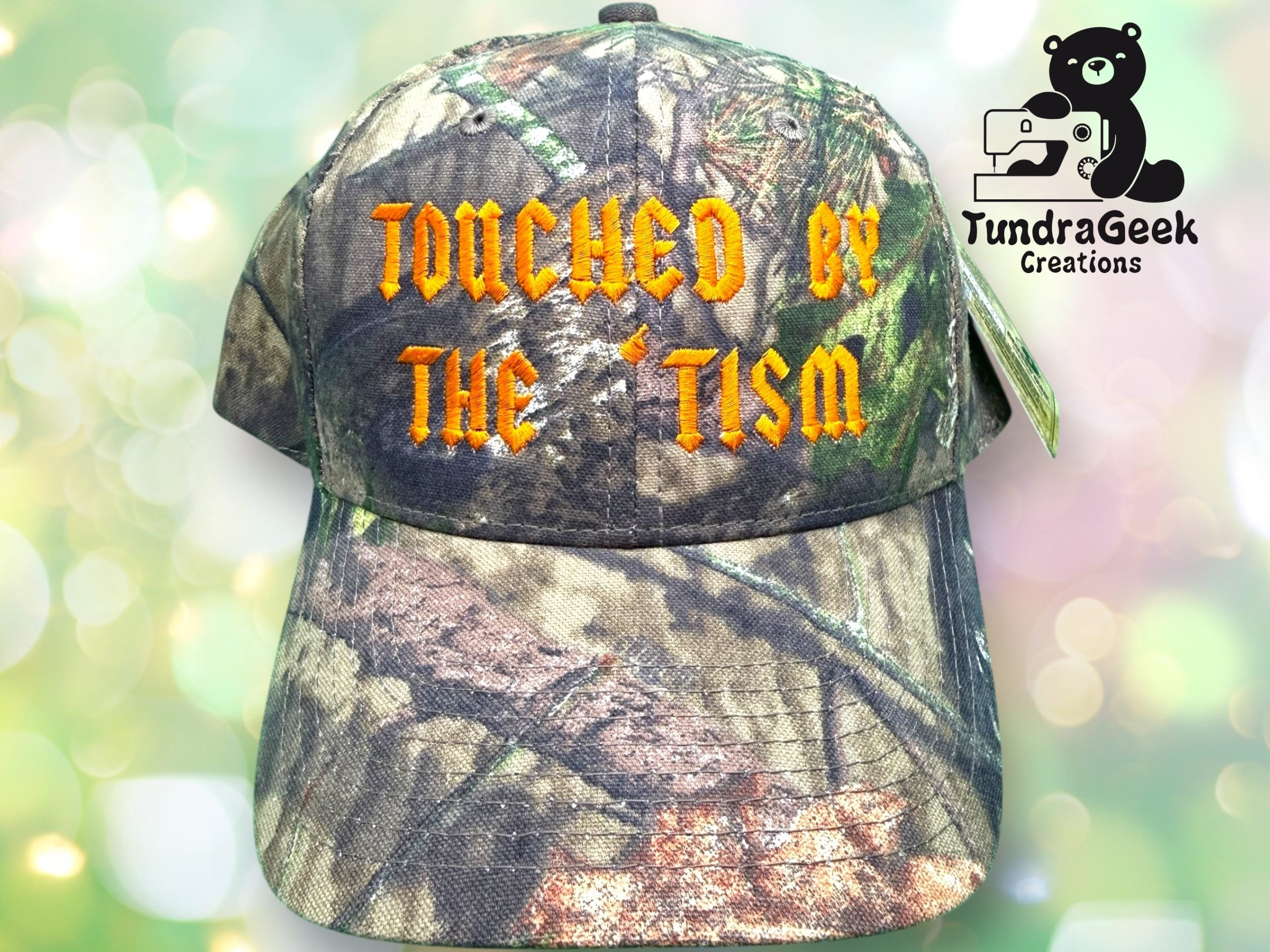 A camouflage baseball cap with yellow-orange text reading TOUCHED BY THE TISM and a TundraGeek Creations logo featuring a bear with a sewing machine.