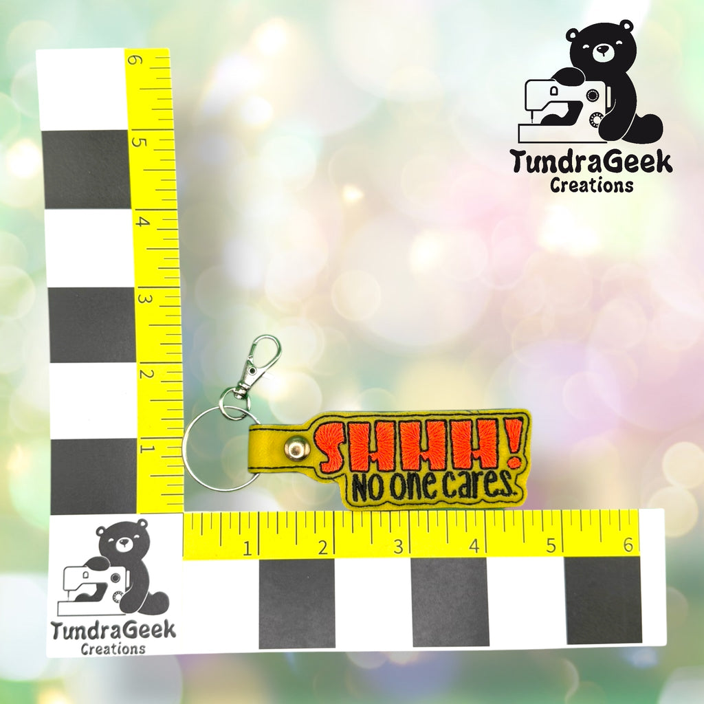 A keychain with a yellow tag reads SHHH! NO ONE CARES in red and yellow letters, placed on a measurement grid with the TundraGeek Creations logo.