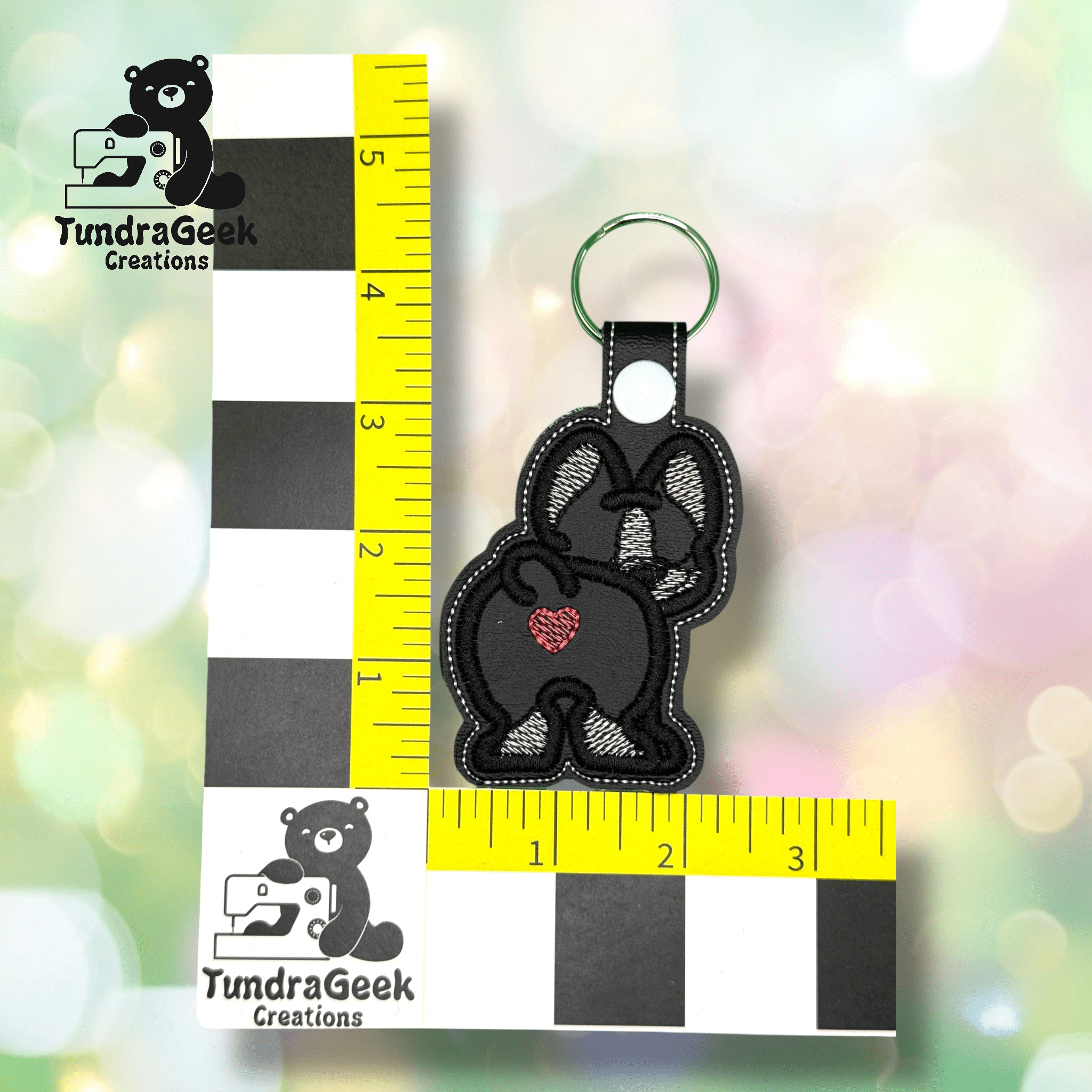 A black and white embroidered keychain shaped like a dogs rear with a red heart, placed next to two measuring rulers, showing it is about 3.5 inches tall.
