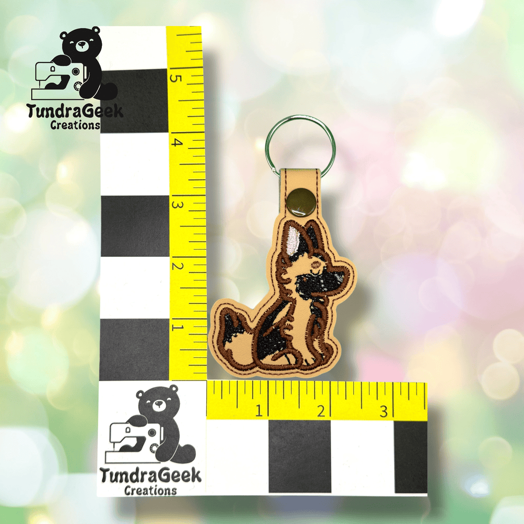 A keychain shaped like a German Shepherd dog next to a ruler for scale, with the TundraGeek Creations logo in the background.