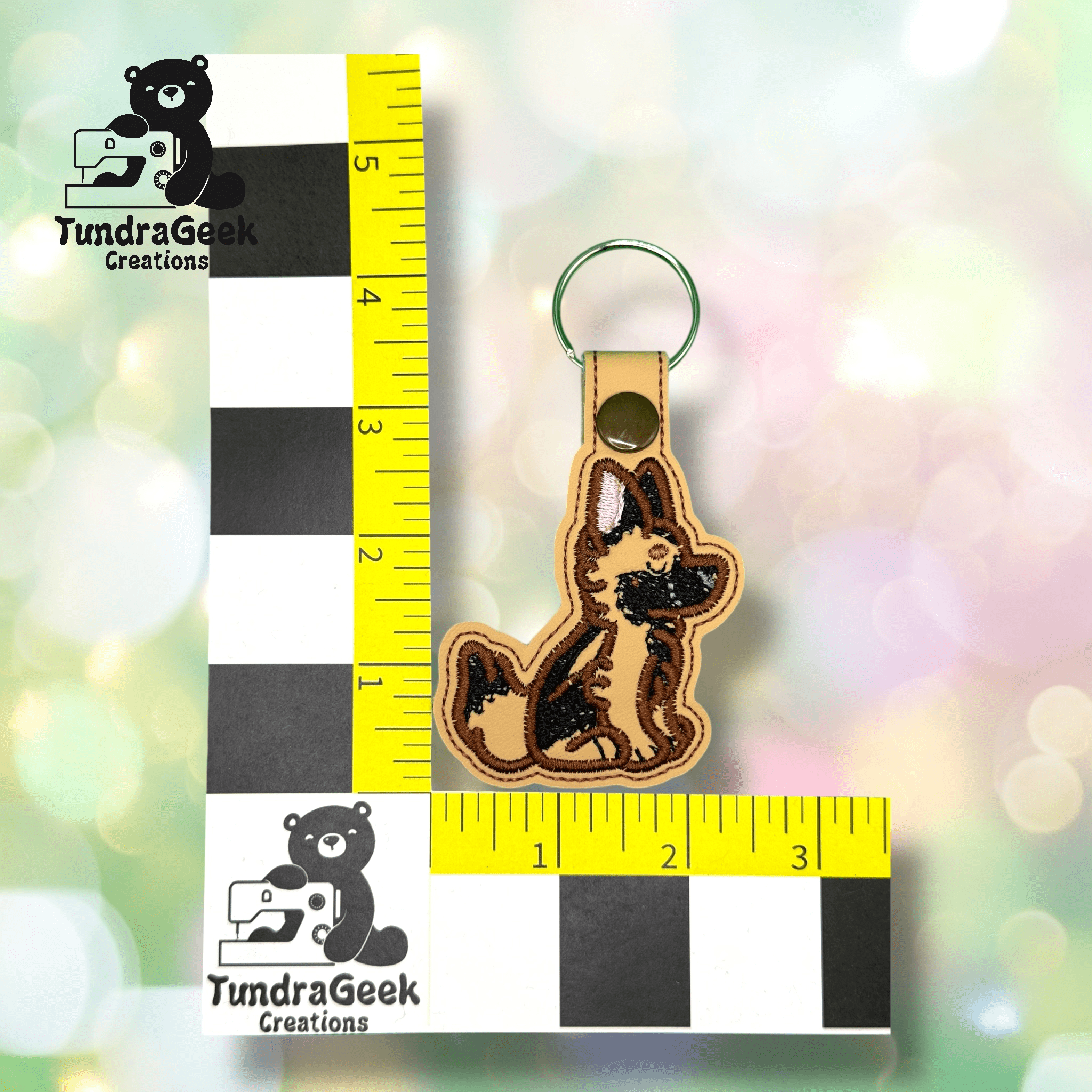 A keychain shaped like a German Shepherd dog next to a ruler for scale, with the TundraGeek Creations logo in the background.