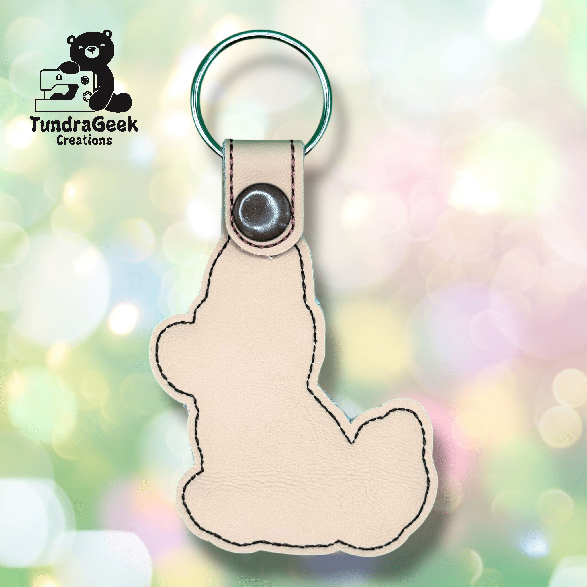 A beige, bear-shaped leather keychain with a snap button and metal ring, displayed against a pastel, bokeh background. TundraGeek Creations logo is visible in the top left corner.