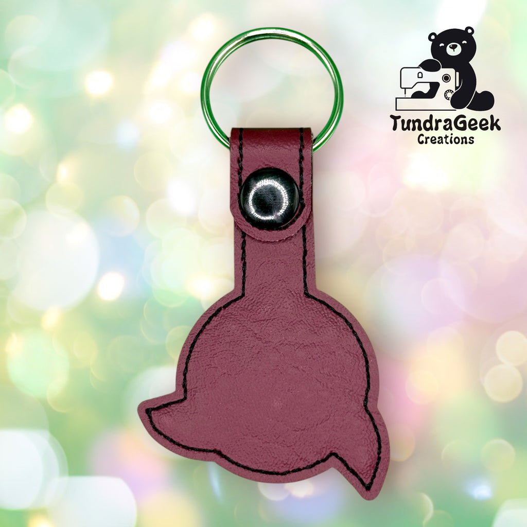 A maroon leather keychain shaped like a whale tail with a metal ring and snap, shown against a blurred light background. The TundraGeek Creations logo is in the top right corner.