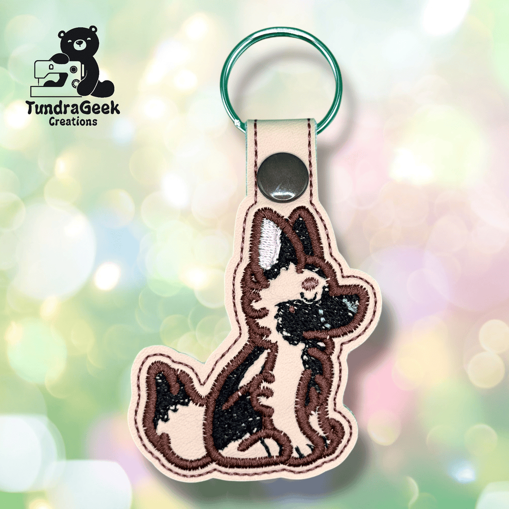 Embroidered keychain featuring a sitting dog with brown, black, and cream coloring, attached to a snap-closed loop and a green keyring. TundraGeek Creations logo in the corner.