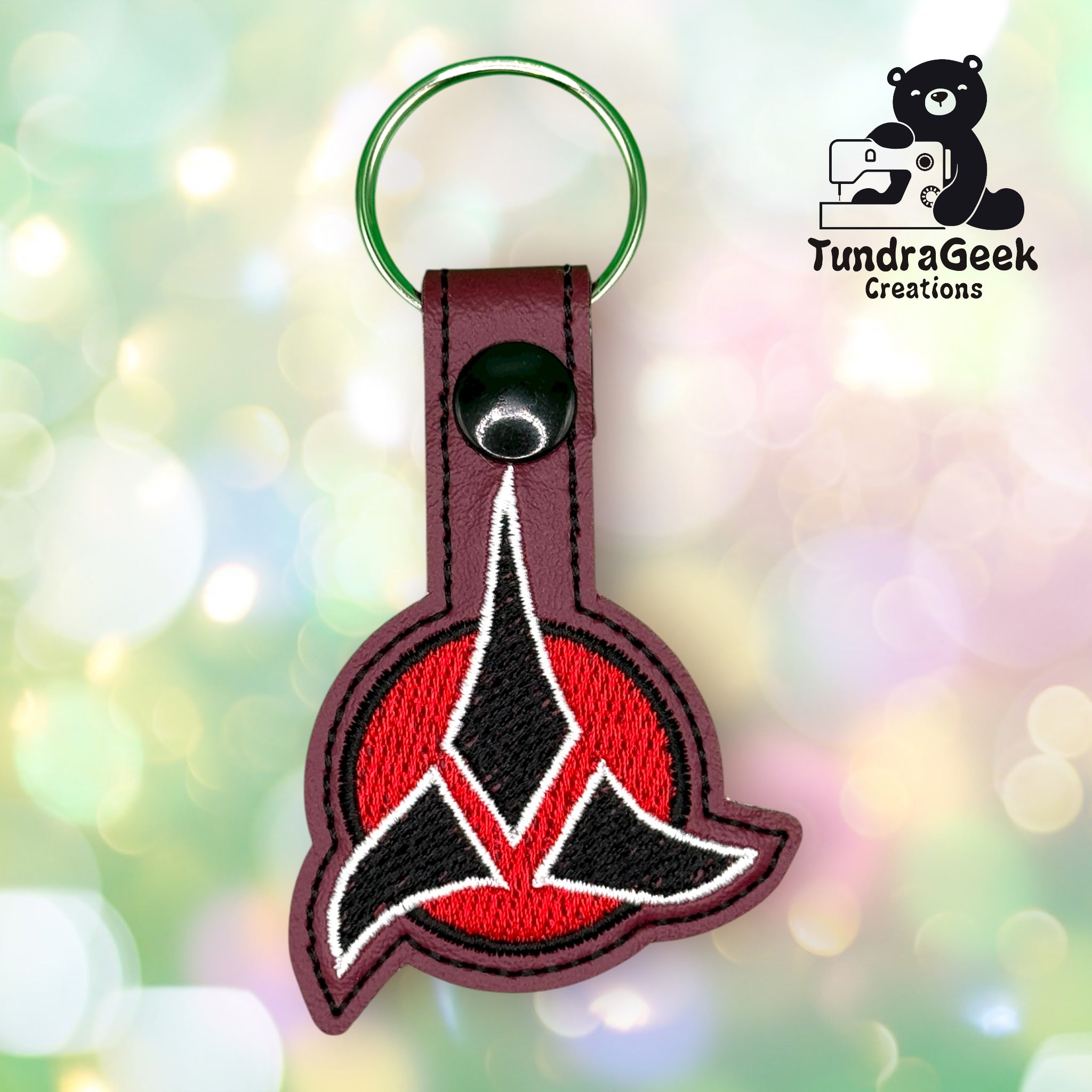 Keychain featuring the Klingon Empire emblem from Star Trek on a red background, made by TundraGeek Creations, shown against a blurred, colorful backdrop.