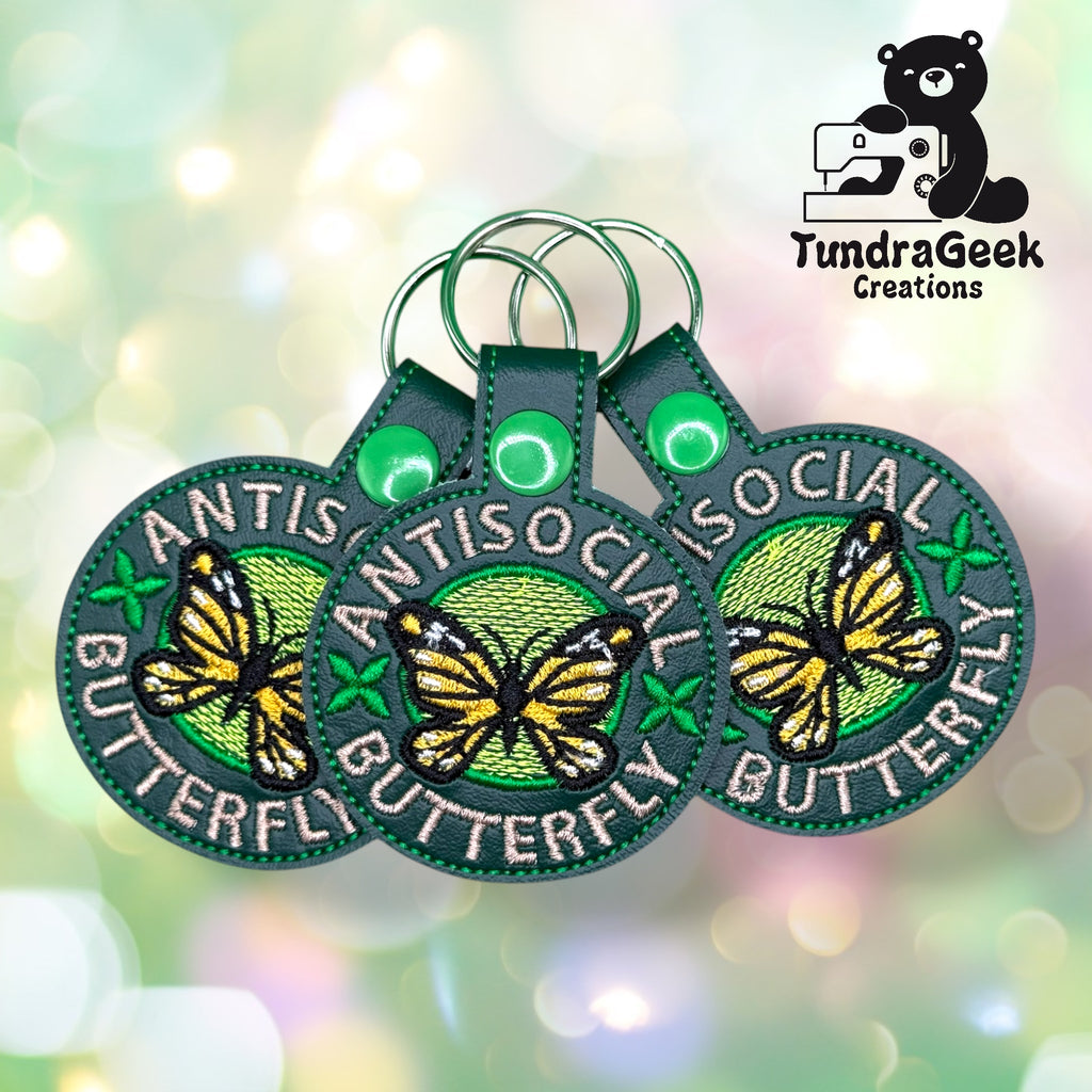 Three embroidered keychains with a yellow butterfly and the words Antisocial Butterfly on green backgrounds, displayed in front of a bokeh-style backdrop with the TundraGeek Creations logo.