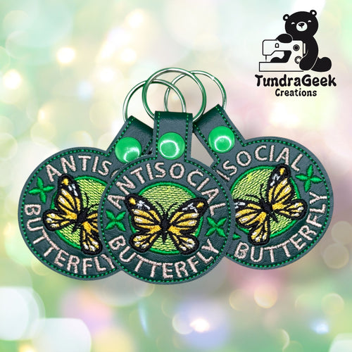 Three embroidered keychains with a yellow butterfly and the words Antisocial Butterfly on green backgrounds, displayed in front of a bokeh-style backdrop with the TundraGeek Creations logo.