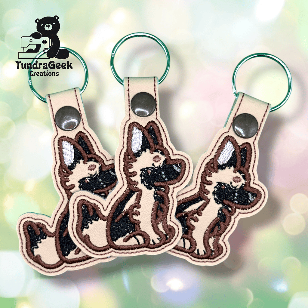 Three embroidered keychains featuring a cartoon German Shepherd dog design, displayed against a soft, pastel, bokeh background. The TundraGeek Creations logo is in the top left corner.