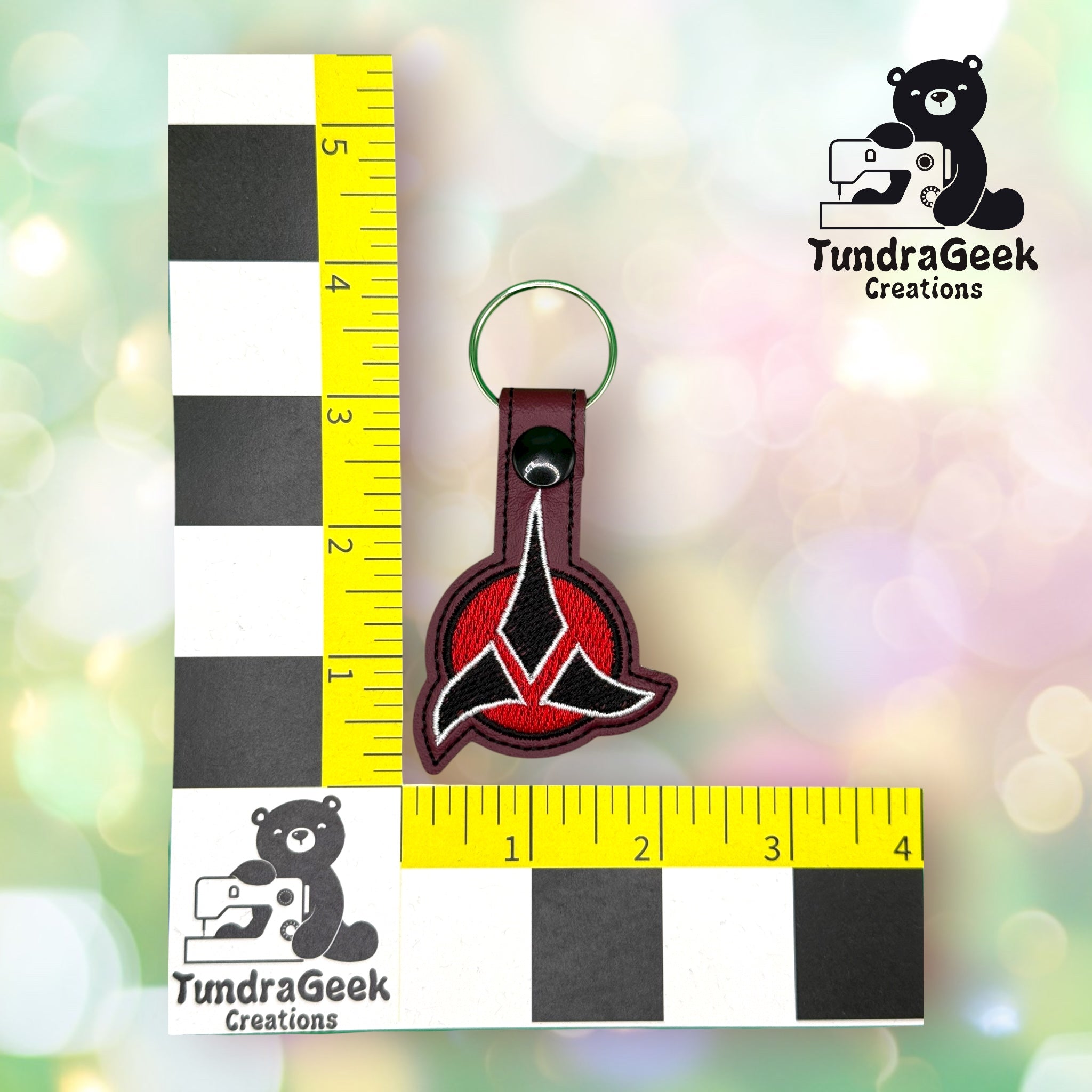 A keychain with a red and black emblem is placed next to a yellow and black ruler showing it is about 3 inches long. The TundraGeek Creations logo appears in two corners.
