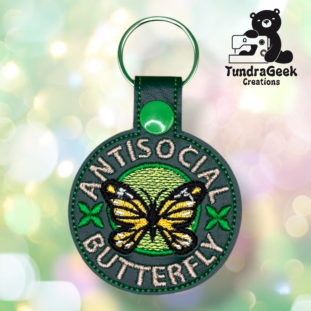 Round embroidered keychain with a butterfly and green accents. Text reads ANTISOCIAL BUTTERFLY. TundraGeek Creations logo with a bear and sewing machine in the top right corner.