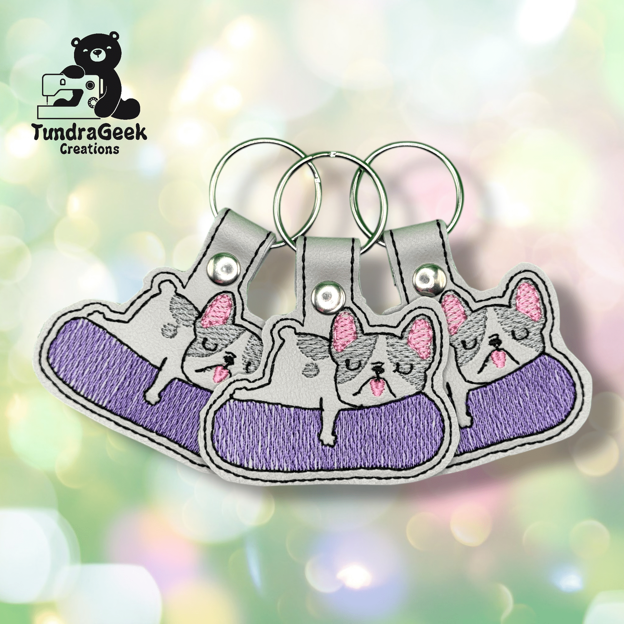 Three embroidered keychains featuring a gray cat lounging on a purple surface, displayed on a soft, pastel bokeh background. TundraGeek Creations logo in the top left corner.