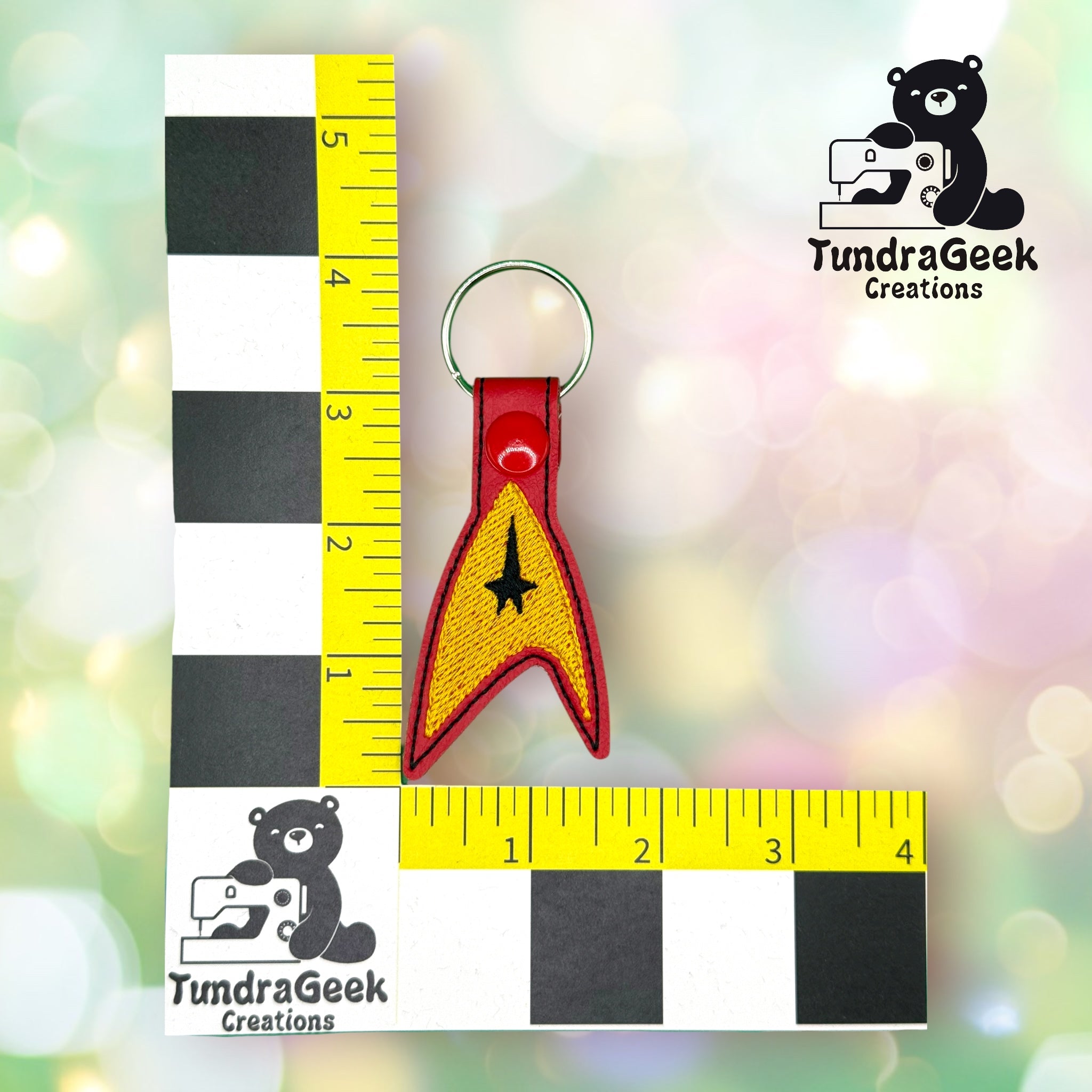 A red and yellow Star Trek insignia keychain, about 3 inches tall, is placed on a measurement grid with the TundraGeek Creations logo.