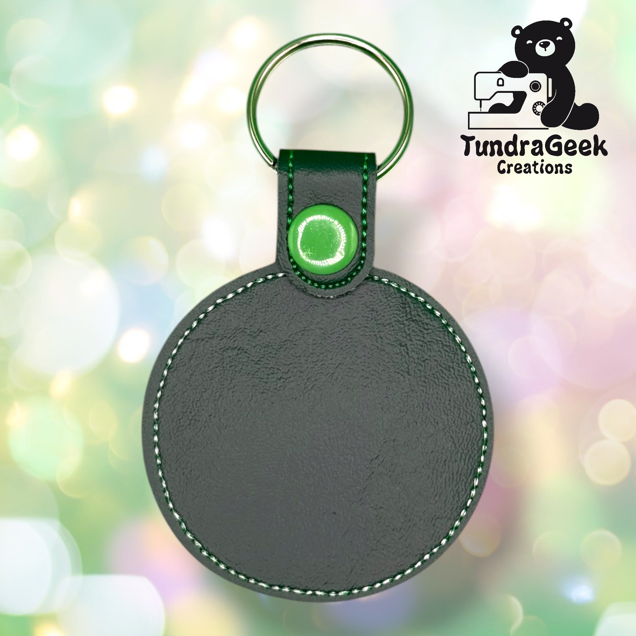 Round dark green leather keychain with green stitching and a metal keyring, displayed against a blurred background with the TundraGeek Creations logo in the corner.