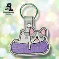 Embroidered keychain featuring a gray and white French Bulldog lying on a purple cushion, with embroidery detail and a metal keyring. TundraGeek Creations logo in the corner.