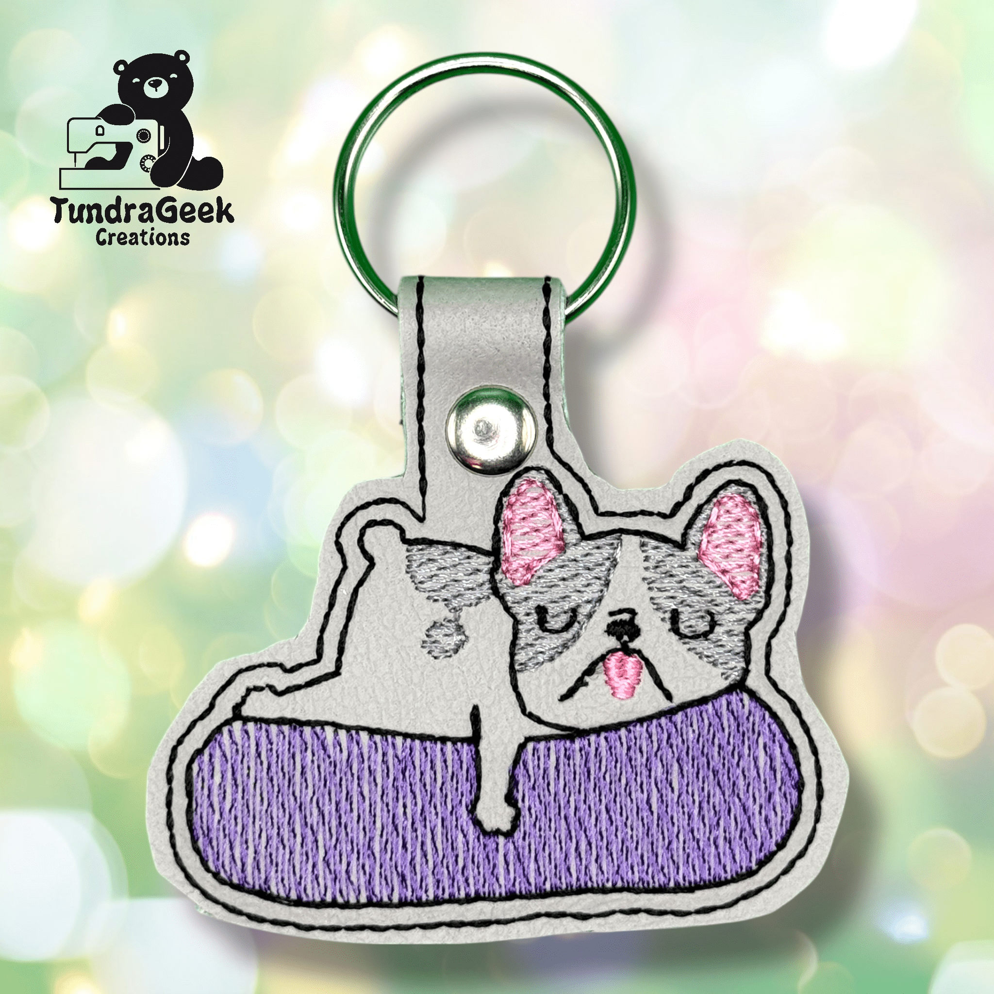Embroidered keychain featuring a gray and white French Bulldog lying on a purple cushion, with embroidery detail and a metal keyring. TundraGeek Creations logo in the corner.