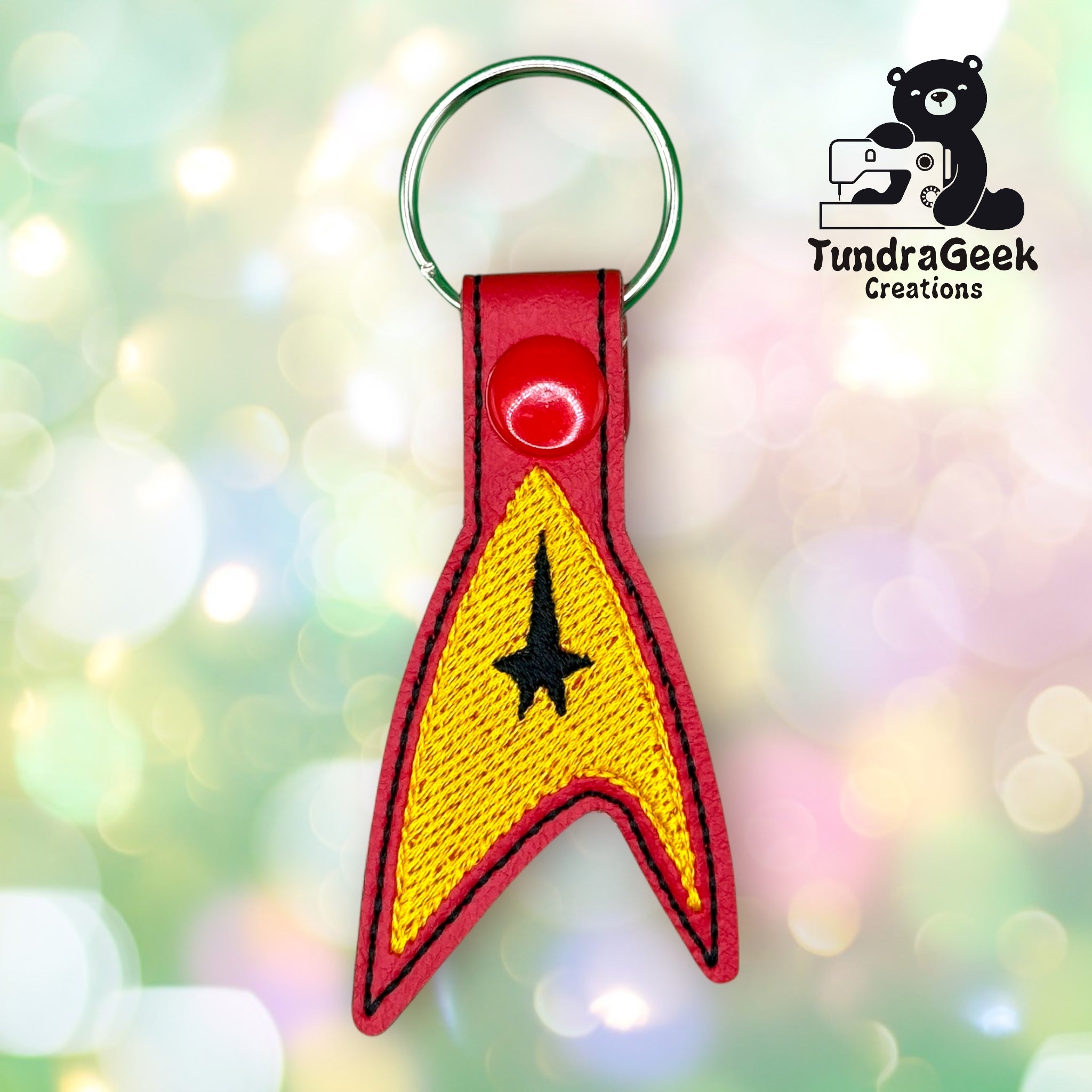 A red and yellow embroidered keychain in the shape of a Star Trek insignia with a black arrow symbol, shown on a colorful blurred background. TundraGeek Creations logo is in the top right.