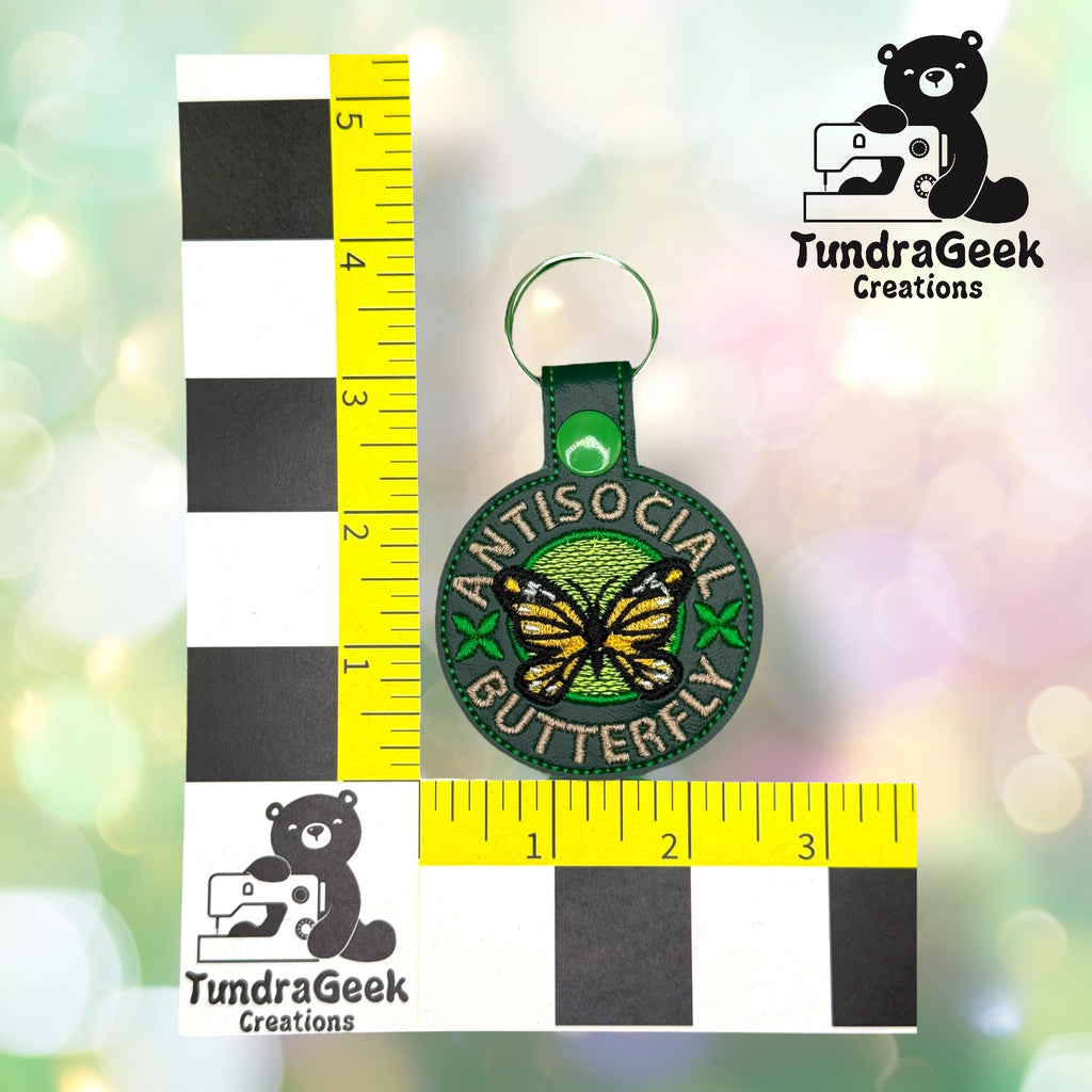 A round embroidered keychain with a butterfly and the text Antisocial Butterfly is shown next to rulers for scale, measuring about 3 inches tall.