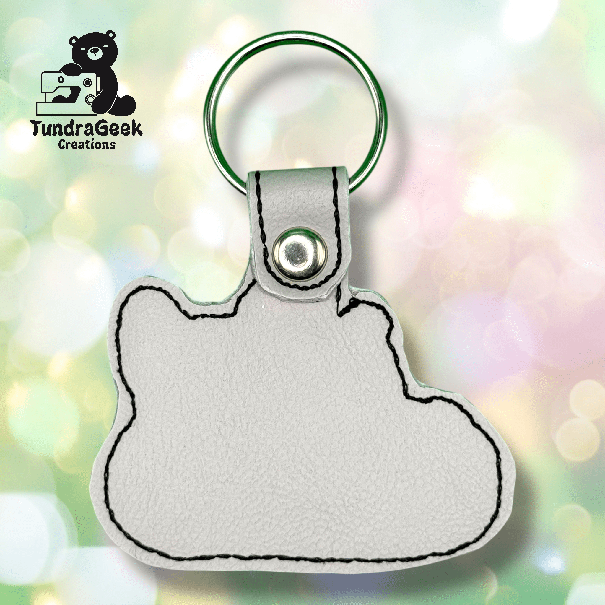 A white faux leather keychain in the shape of a bear head, with black stitching and a metal keyring, shown on a pastel bokeh background. TundraGeek Creations logo in the corner.