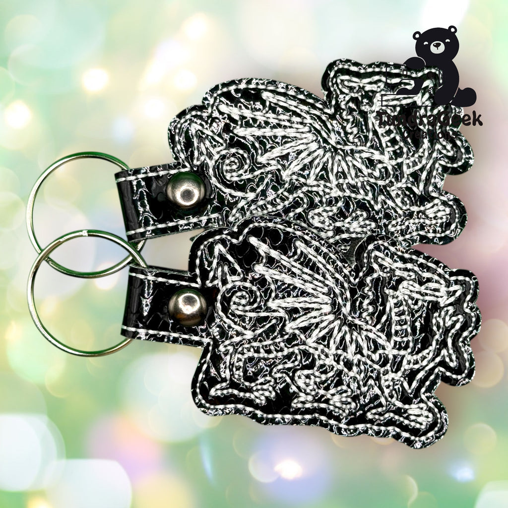 Two embroidered keychains with intricate dragon designs on black material, each attached to a metal key ring, are displayed against a blurred, pastel background with a bear logo in the corner.