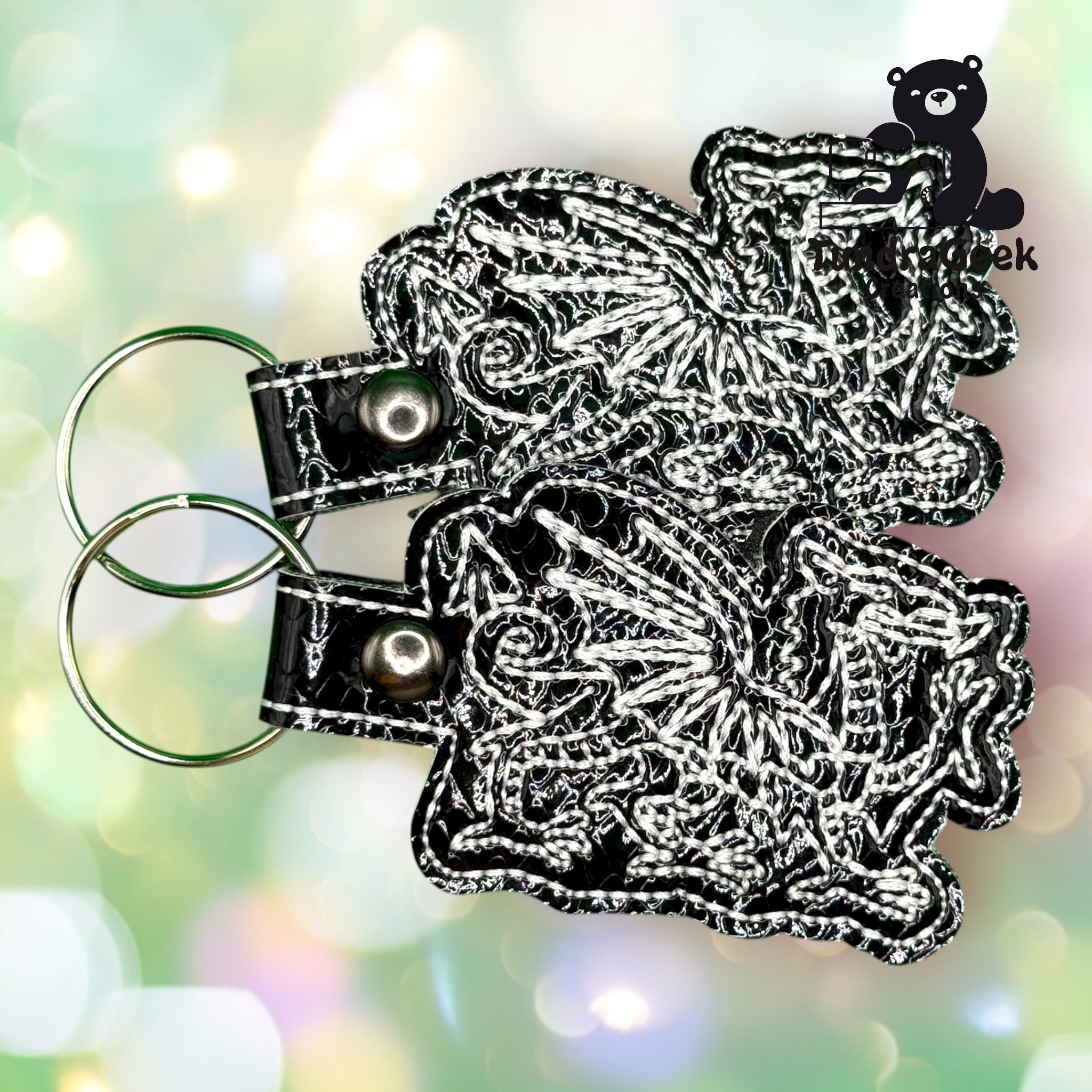 Two embroidered keychains with intricate dragon designs on black material, each attached to a metal key ring, are displayed against a blurred, pastel background with a bear logo in the corner.