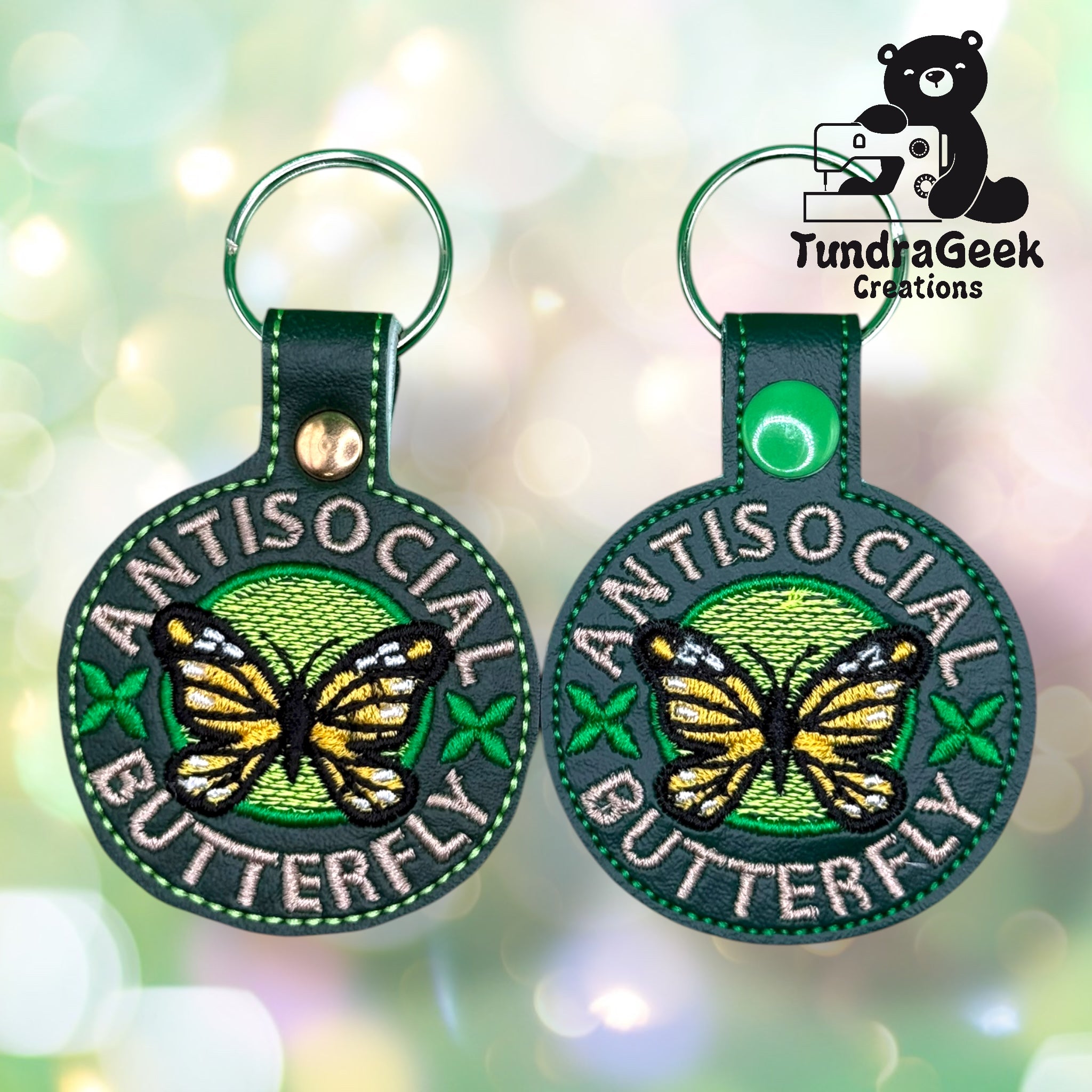 Two round keychains with embroidered yellow butterflies, green backgrounds, and the words ANTISOCIAL BUTTERFLY. Logo for TundraGeek Creations is in the upper right corner.