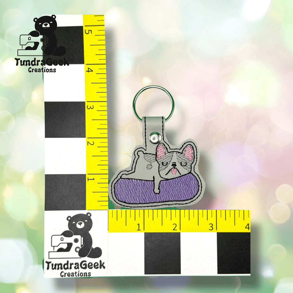 Embroidered keychain of a gray French bulldog lying on a purple cushion, shown next to a ruler for scale, measuring about 3 inches tall.