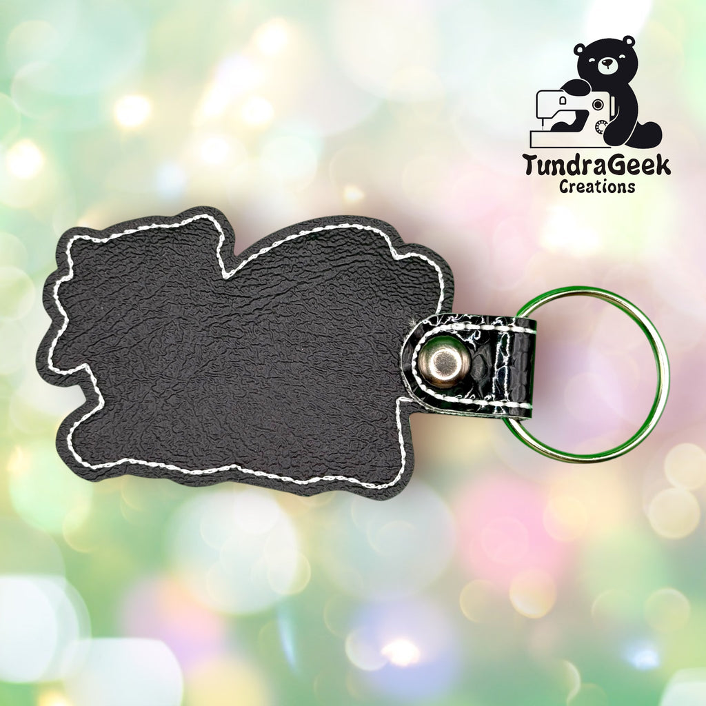 A black faux leather keychain with white stitching and a metal ring, shown against a soft, colorful bokeh background. TundraGeek Creations logo is in the top right corner.