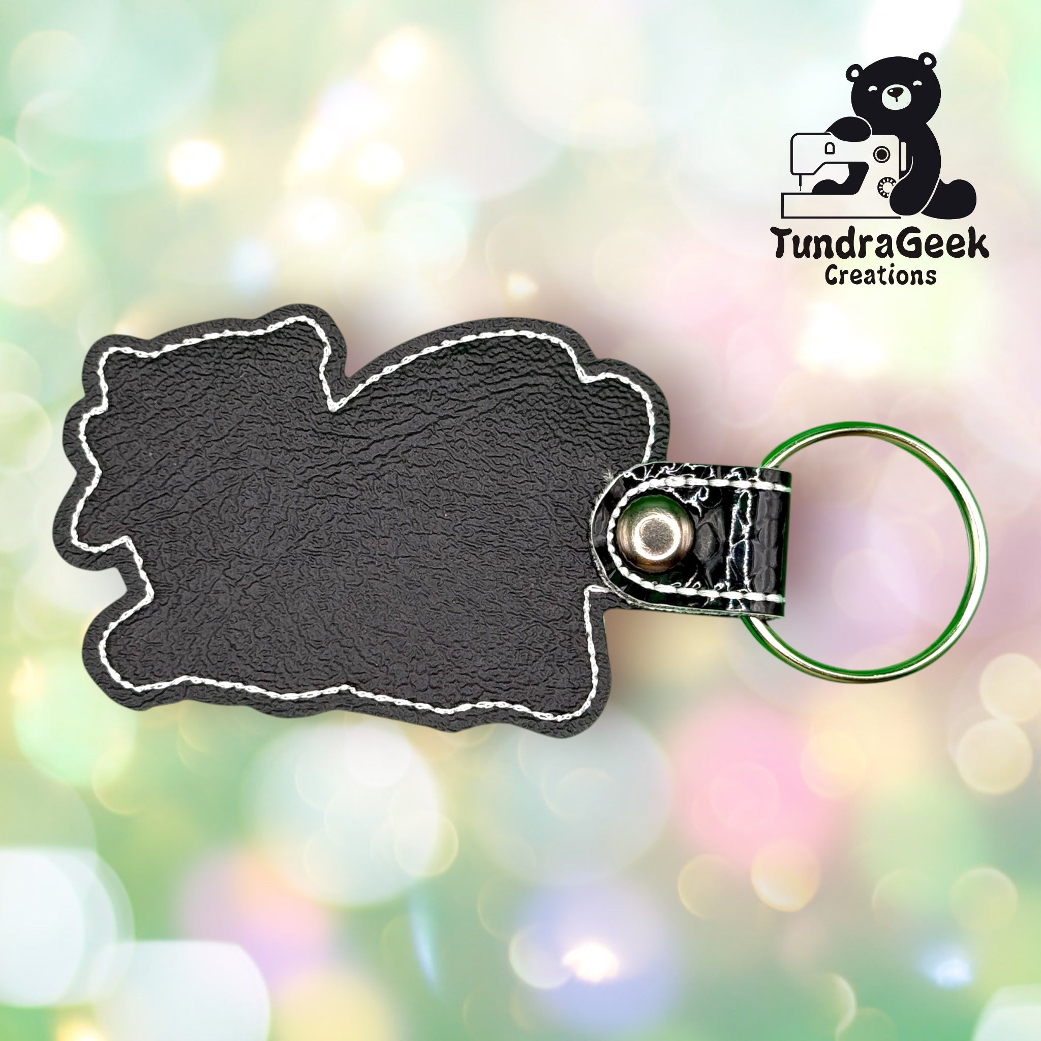 A black faux leather keychain with white stitching and a metal ring, shown against a soft, colorful bokeh background. TundraGeek Creations logo is in the top right corner.