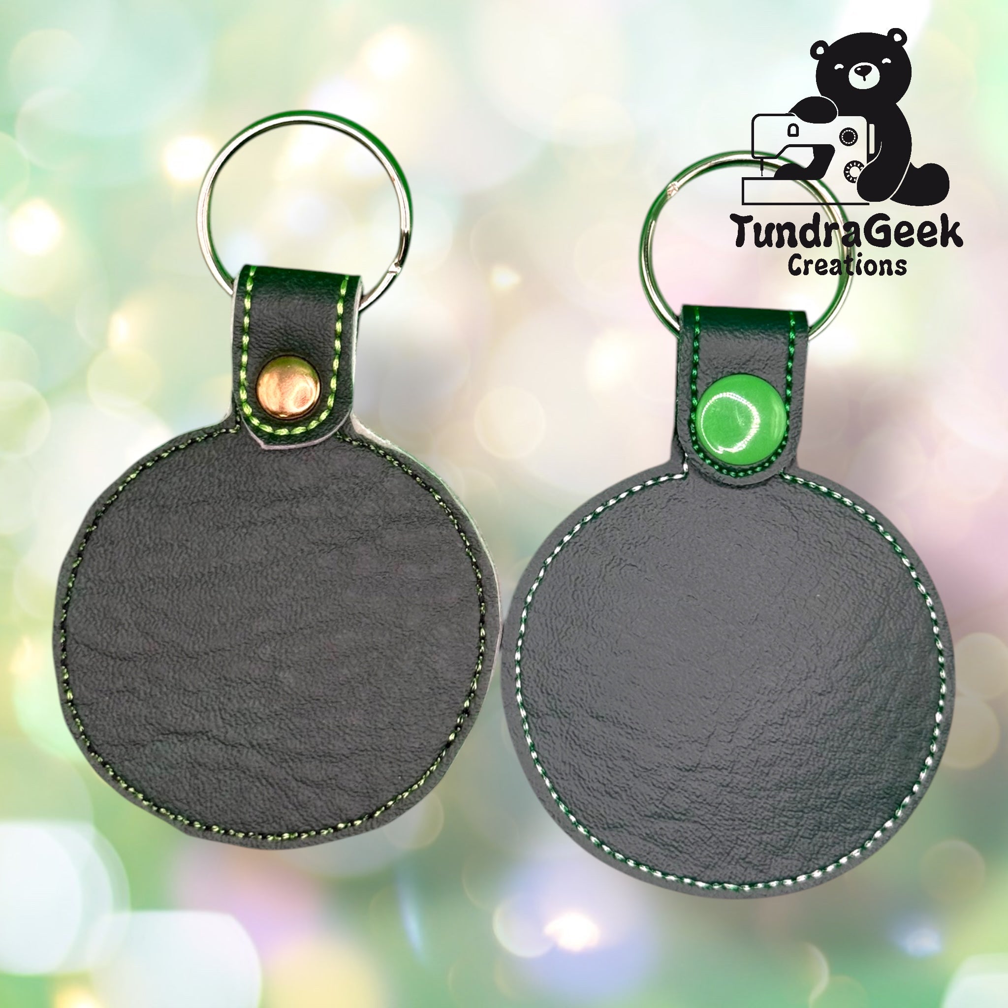 Two circular black leather keychains with green stitching and button snaps, displayed side by side on a blurred light background. TundraGeek Creations logo in upper right corner.