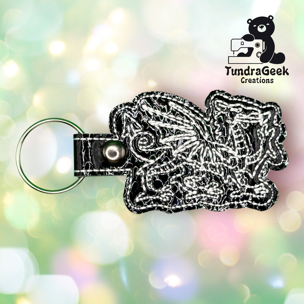 A black and white embroidered dragon keychain with a metal ring, displayed in front of a colorful bokeh background with the TundraGeek Creations logo.