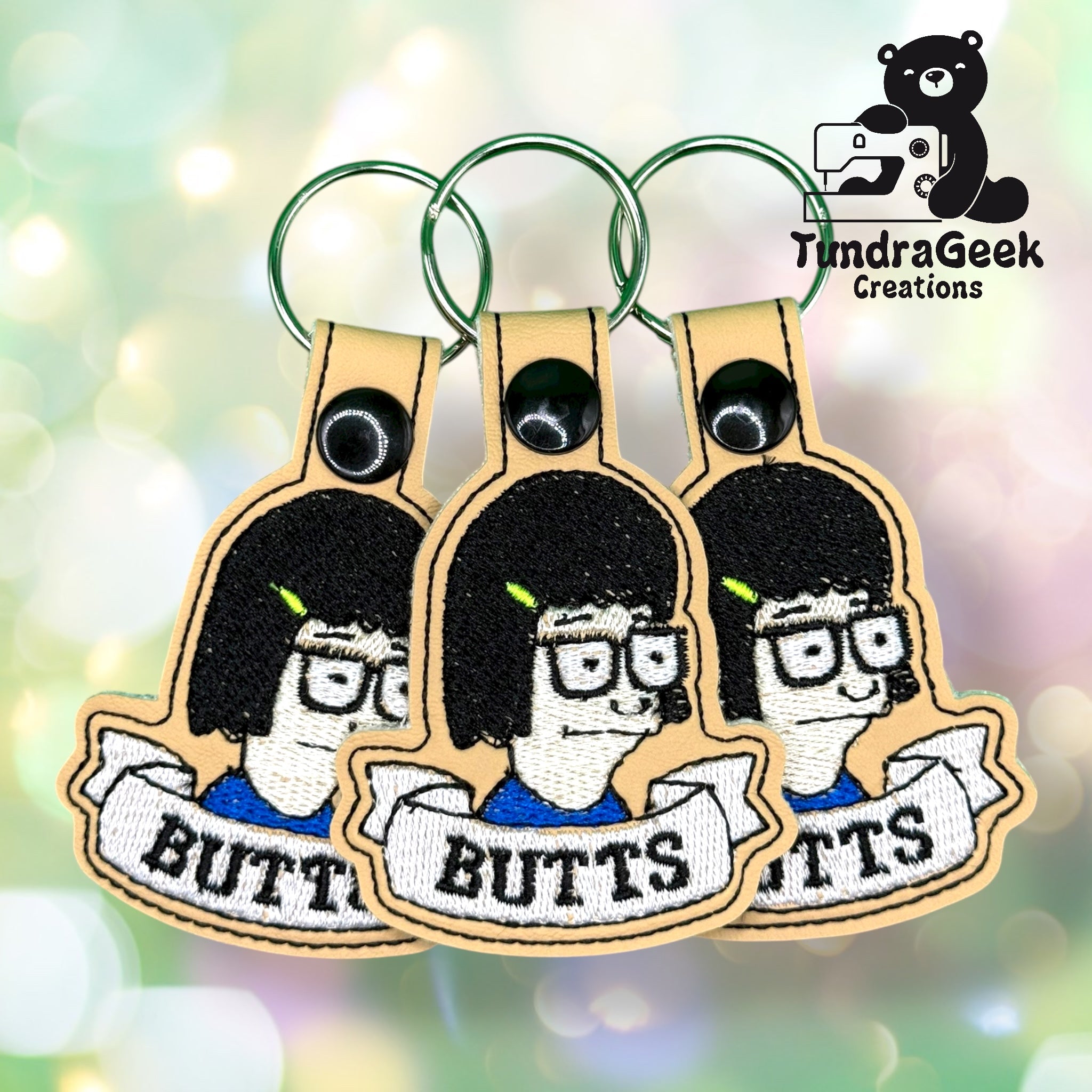 Three embroidered keychains featuring a cartoon character with glasses and short black hair above a banner labeled BUTTS. TimdraGeek Creations logo is in the top right corner.