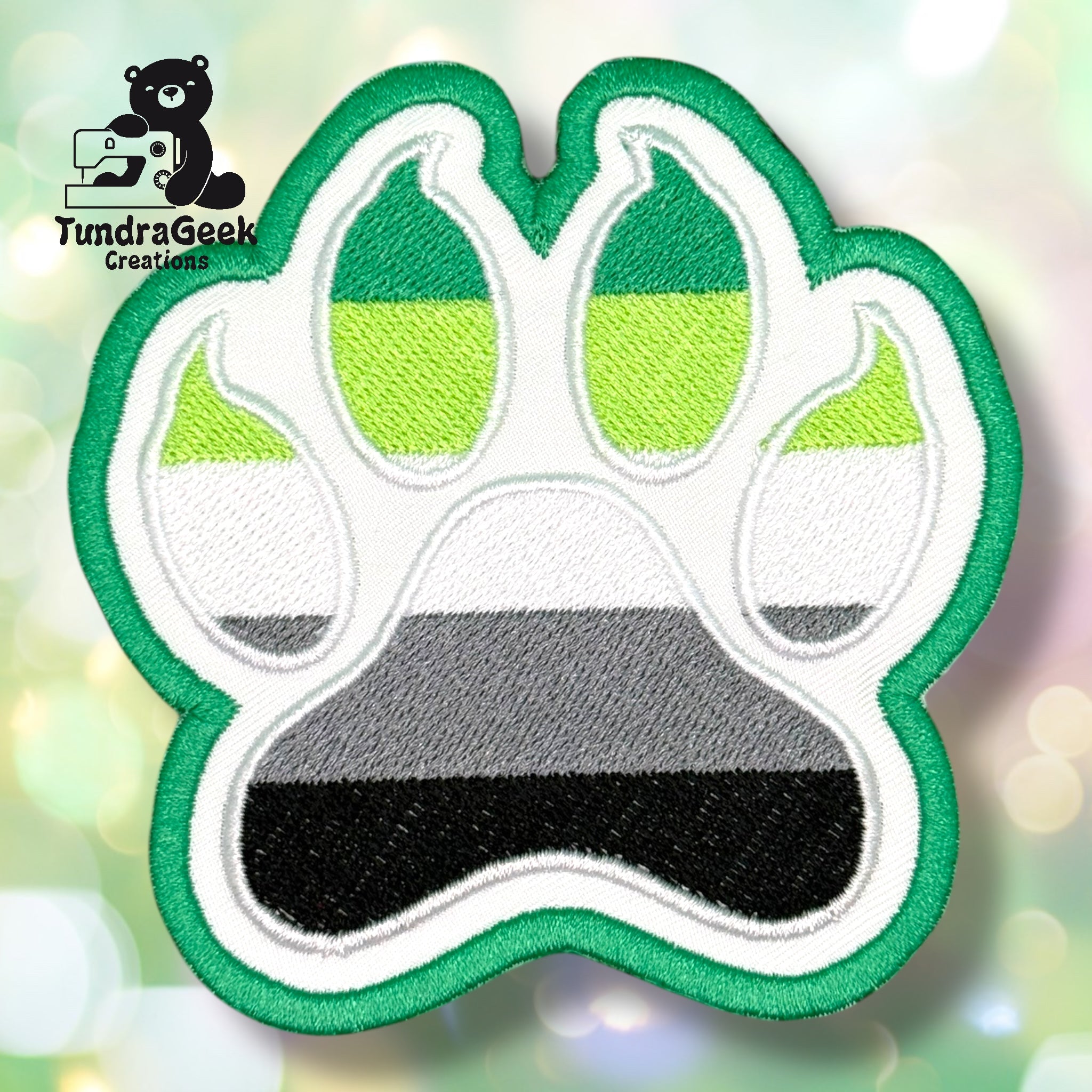 An embroidered paw-shaped patch in aromantic pride flag colors — dark green, light green, white, gray, and black horizontal stripes. The patch features a rounded paw shape with inner pad detail outlined in white. Dark green border edging