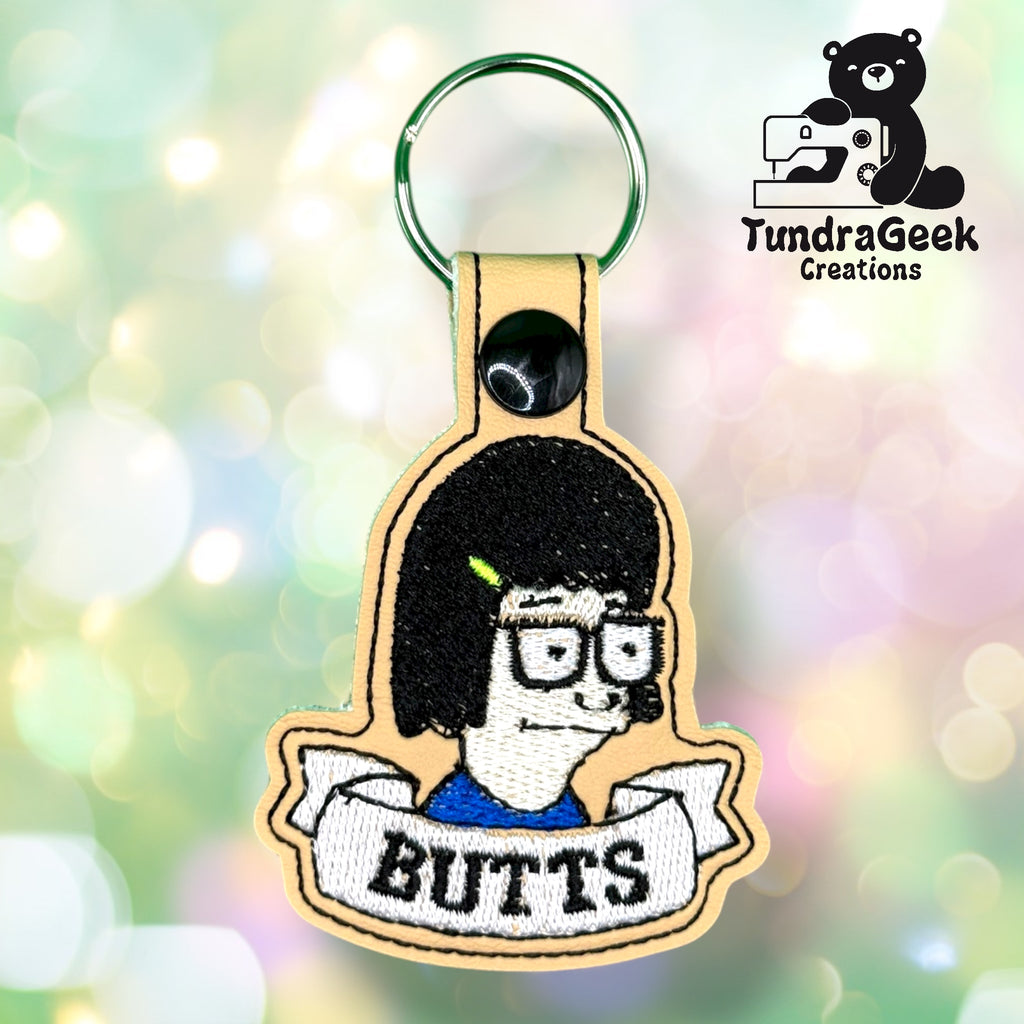 Embroidered keychain featuring a cartoon character with glasses and dark hair above a banner that reads BUTTS. Logo for TundraGeek Creations in the top right corner.