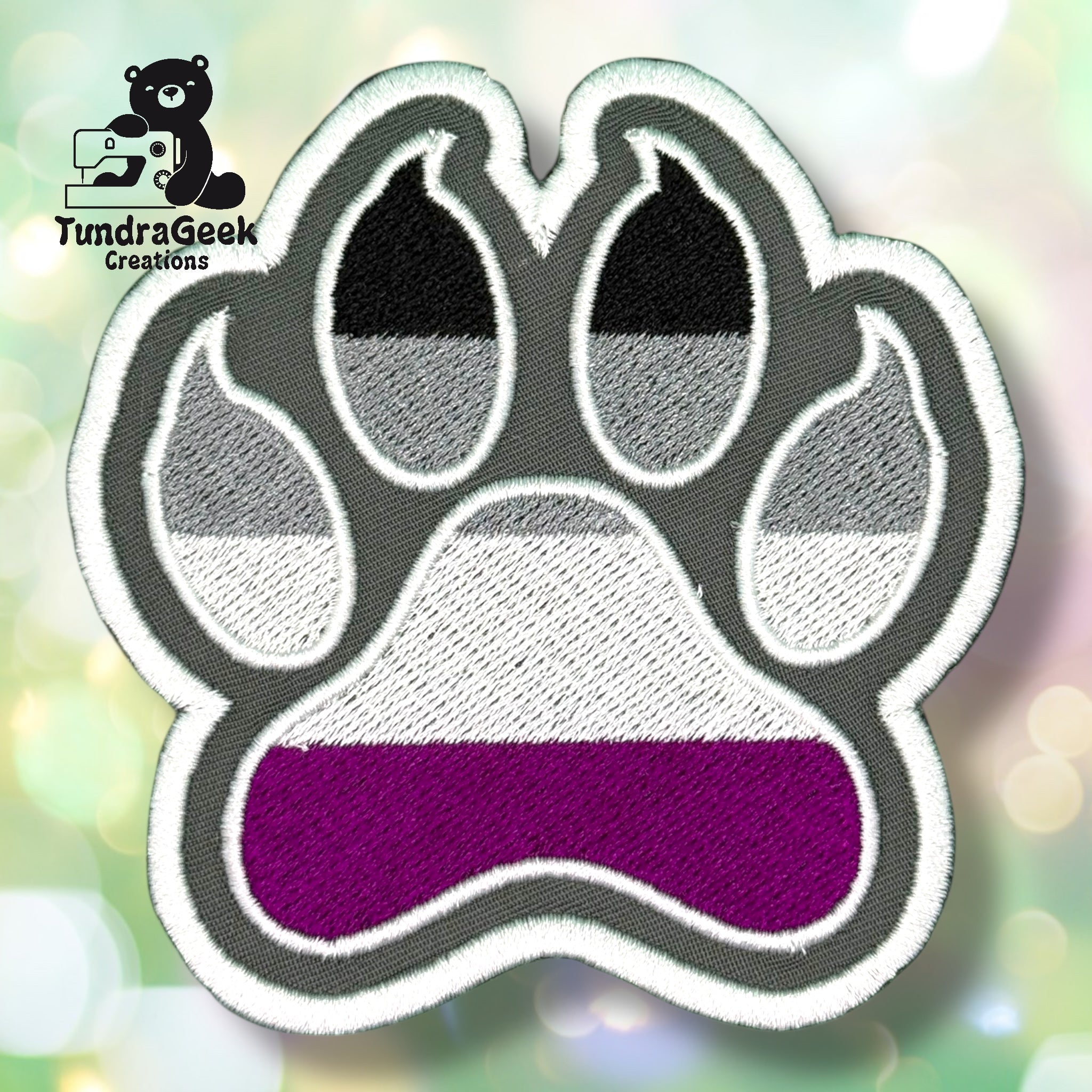An embroidered paw-shaped patch in asexual pride flag colors — horizontal stripes of black, gray, white, and purple. The paw features a rounded shape with inner pad and toe bean details outlined in white. Gray background with white border edging.