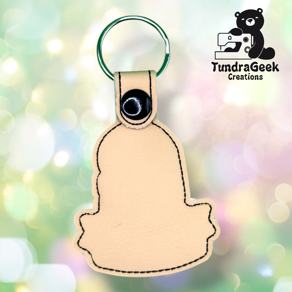 Beige leather keychain in a stylized animal shape with a snap button and metal keyring, displayed against a light, bokeh background. TundraGeek Creations logo in the top right corner.