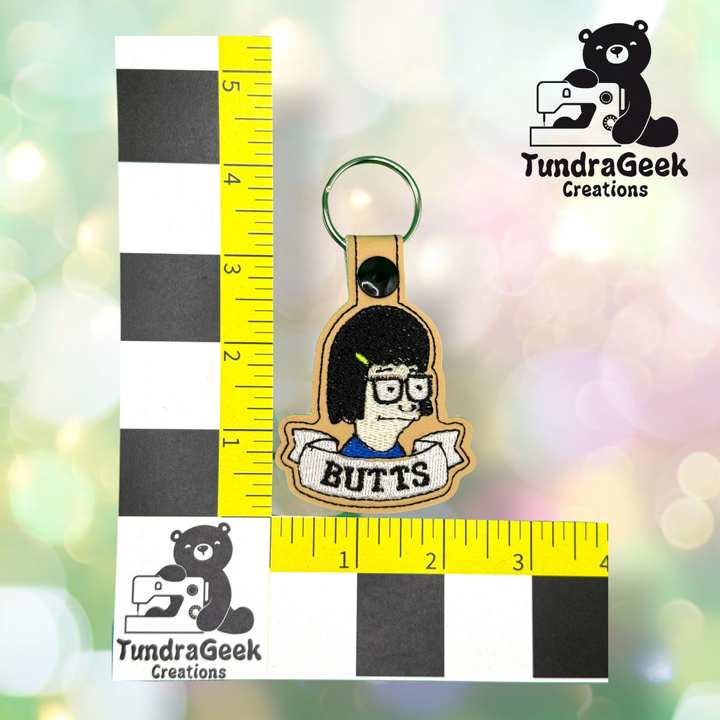 Embroidered keychain with cartoon character, glasses, and BUTTS text. Measured with rulers; TundraGeek Creations bear logo present.